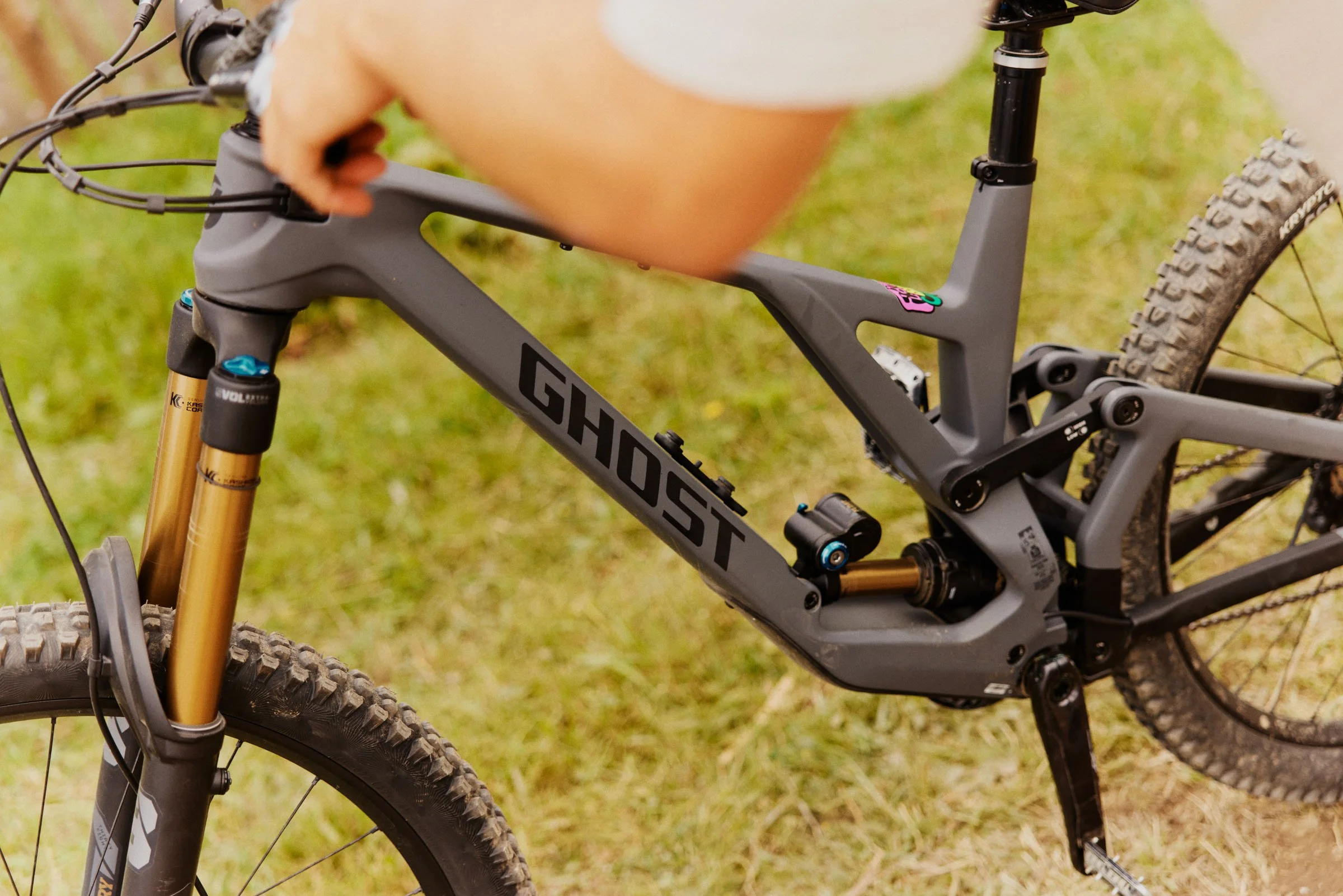 Close-up of a gray mountain bike with the word 'GHOST' on the frame, showing the front suspension fork, rear suspension, and part of the tires, on a grassy area.