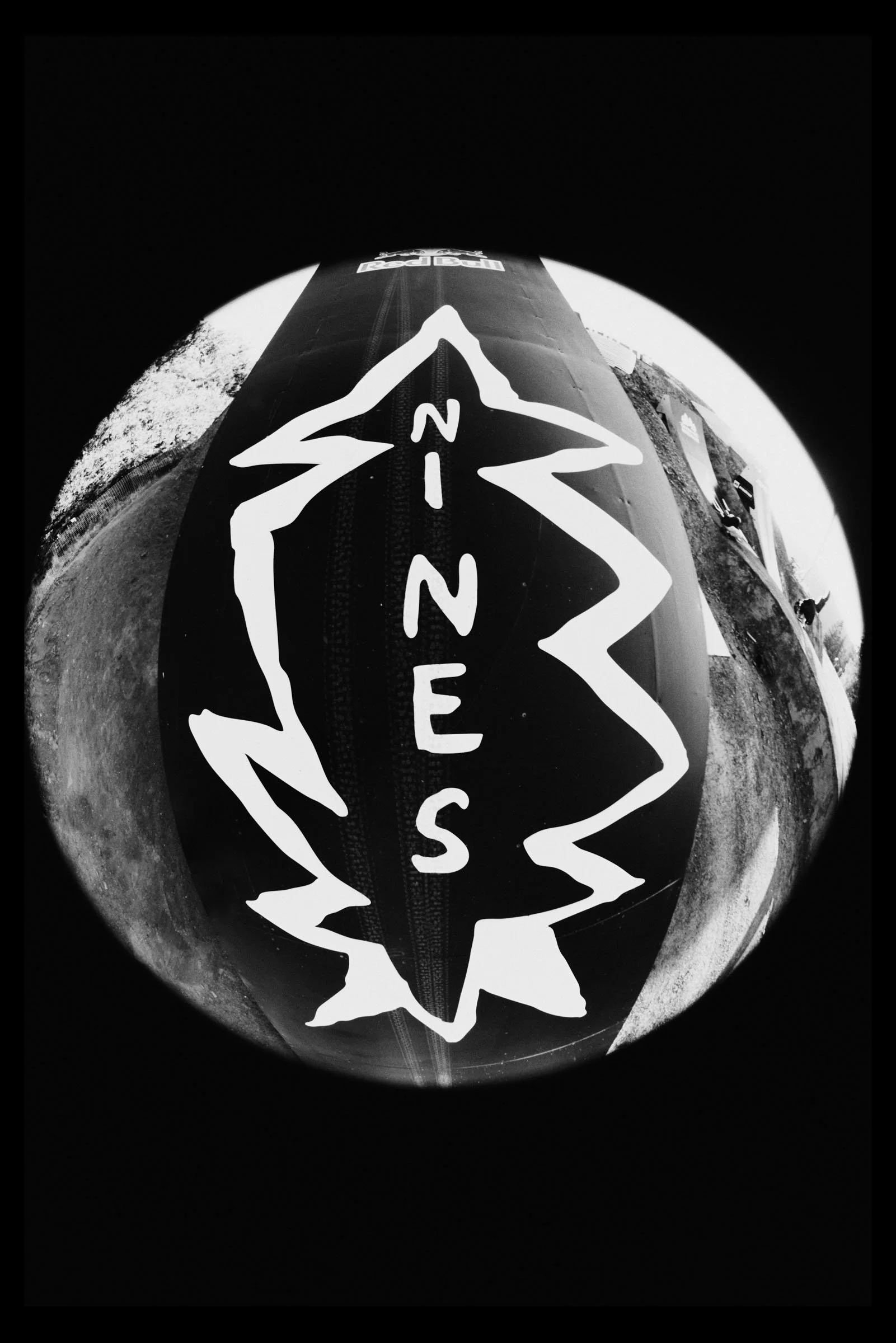 A black and white, fisheye photo of a snowboard with a logo that reads 'NIES' in the center, placed on the snow with trees and sky in the background.
