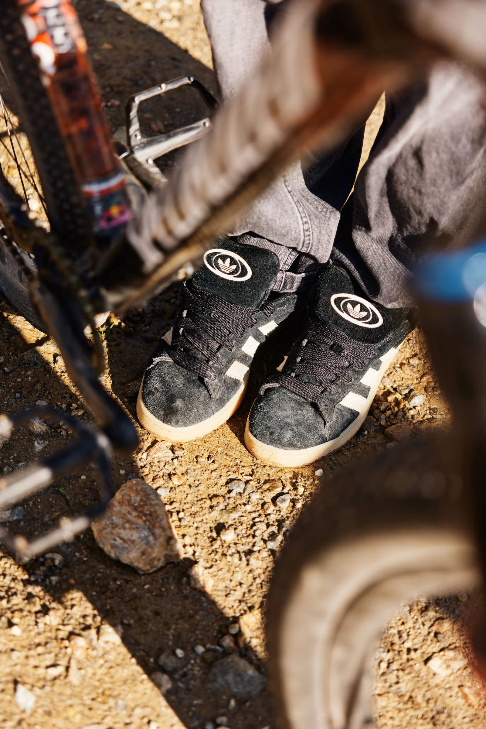 Close-up of a pair of black sneakers with white stripes and a white logo on top, worn by someone standing on rocky, dirt terrain near a bicycle.