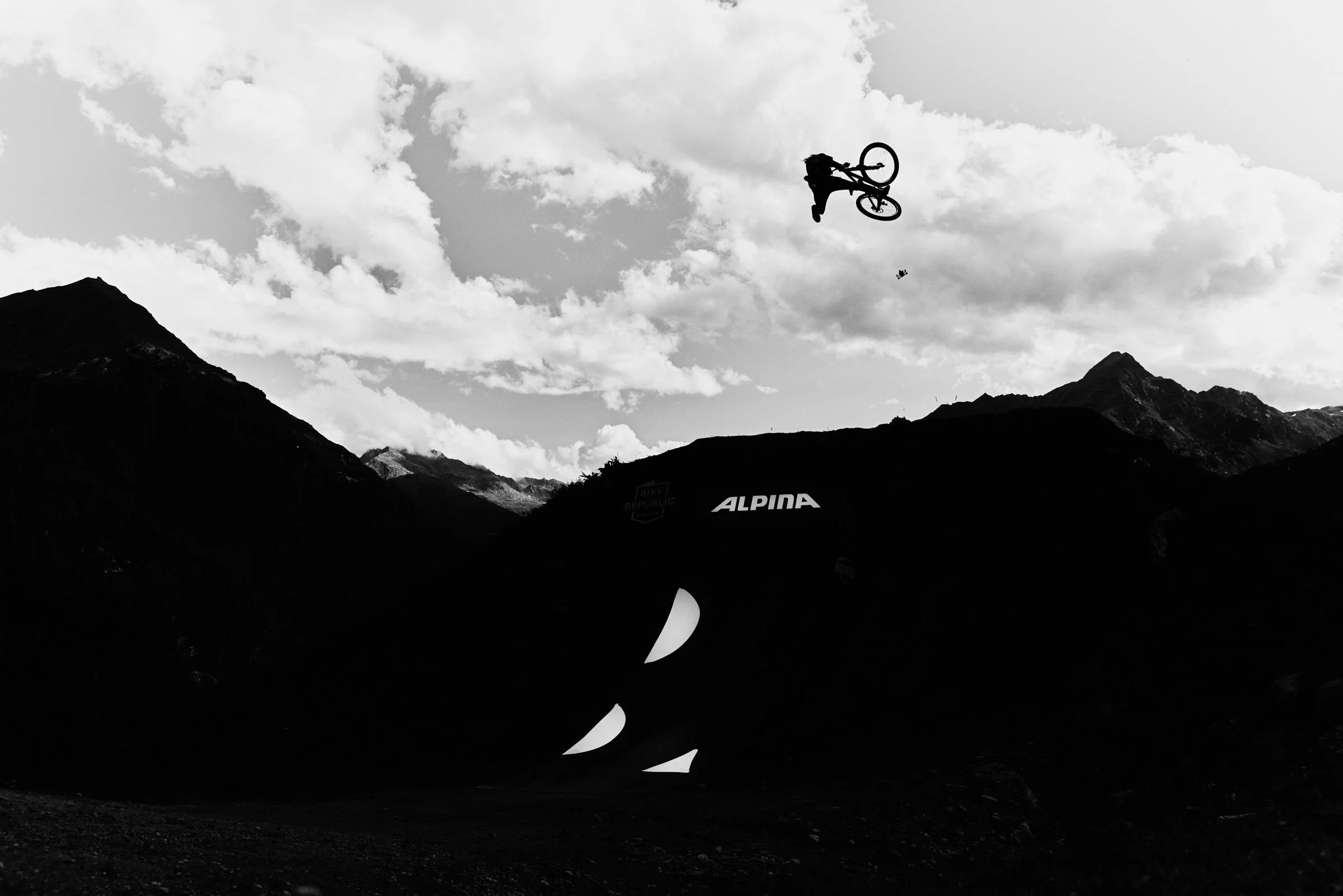 Lukas Schafer Alpina. A silhouette of a mountain biker in mid-air performing a trick against a mountain landscape with clouds in the sky, in black and white.