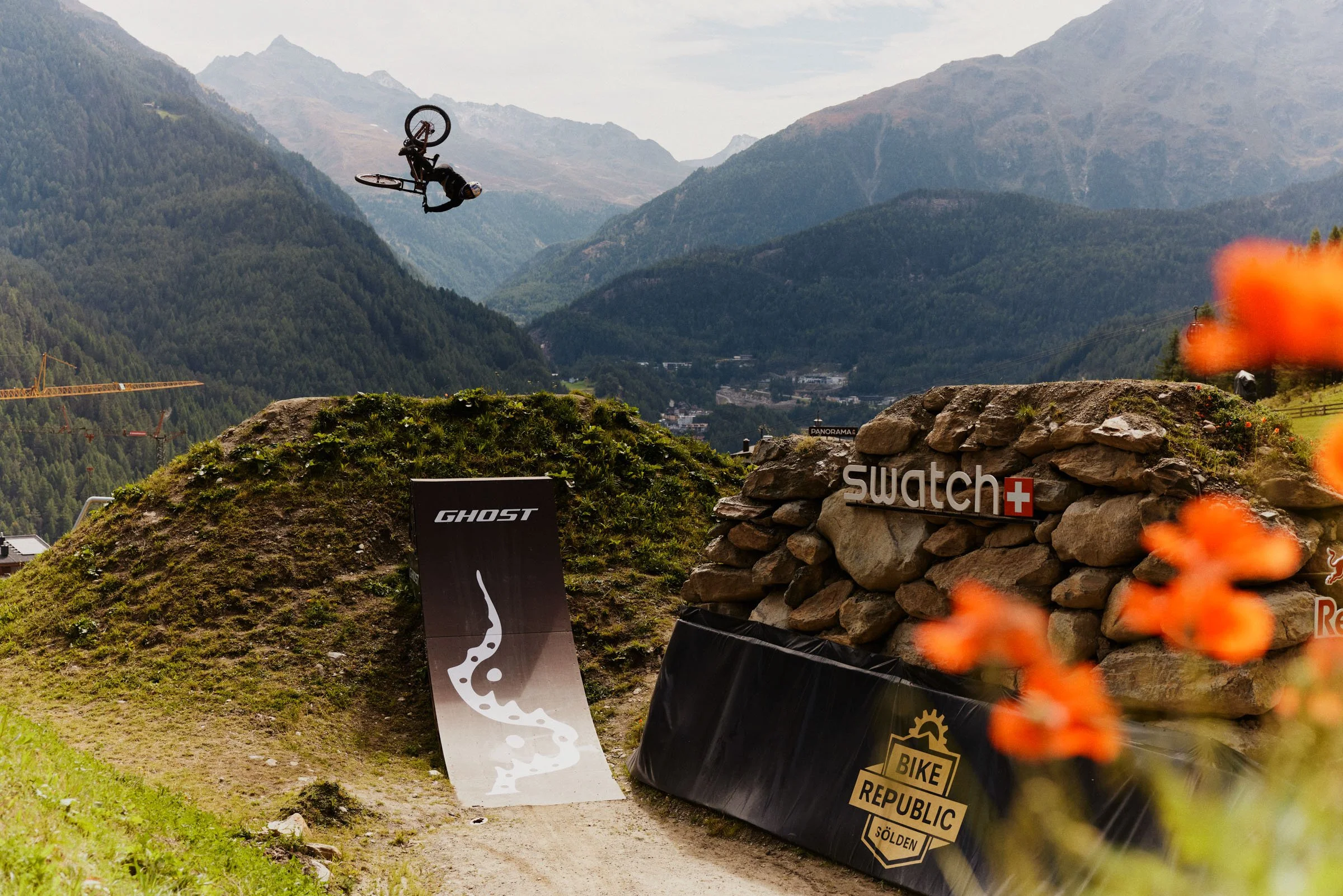 A mountain biking stunt with a rider mid-air over a dirt jump, mountain landscape in the background, with branded banners for Swatch and Ghost, and orange flowers in the foreground.