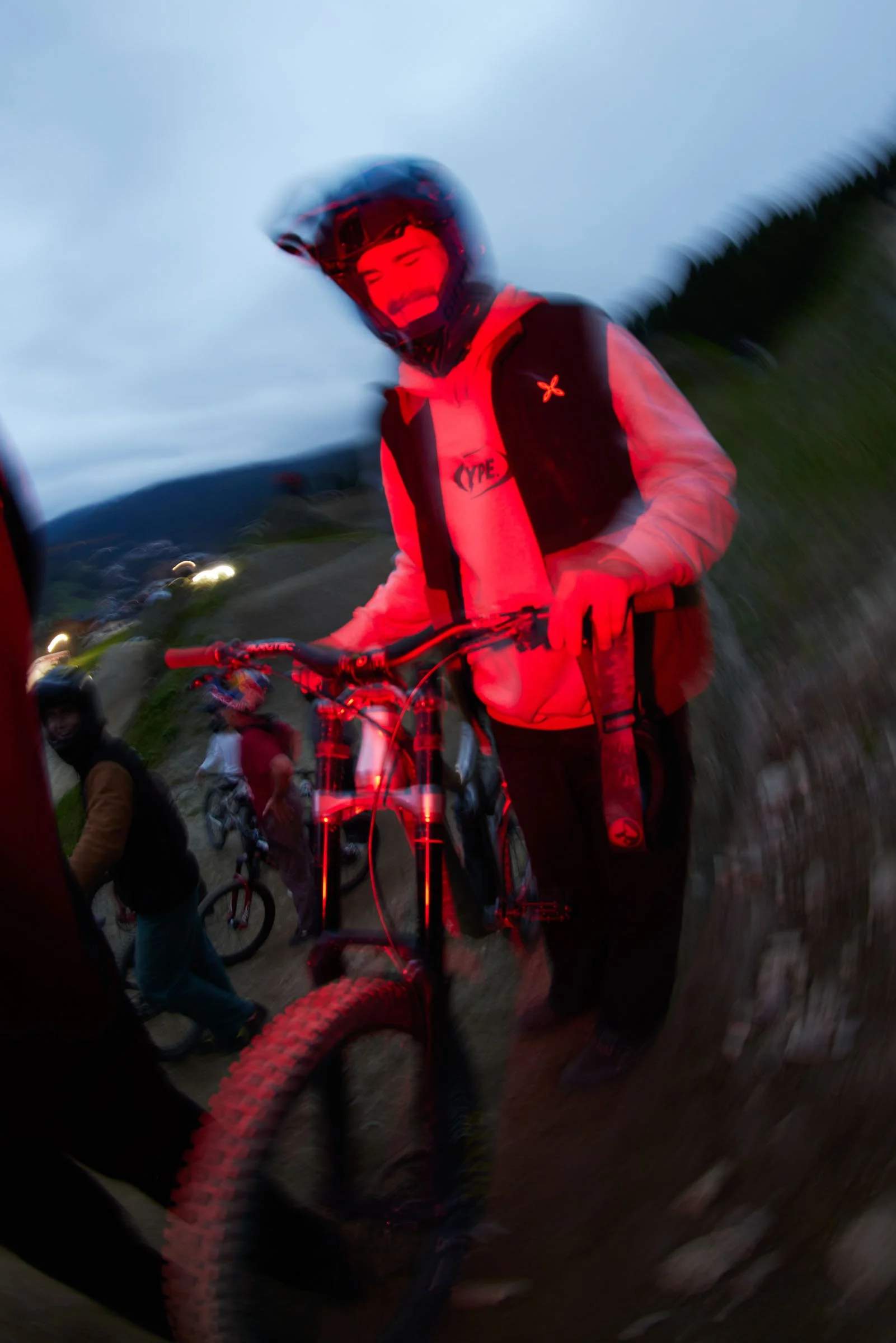 A person wearing a helmet and holding a mountain bike, standing on a dirt trail at dusk. Multiple other cyclists are visible in the background.
