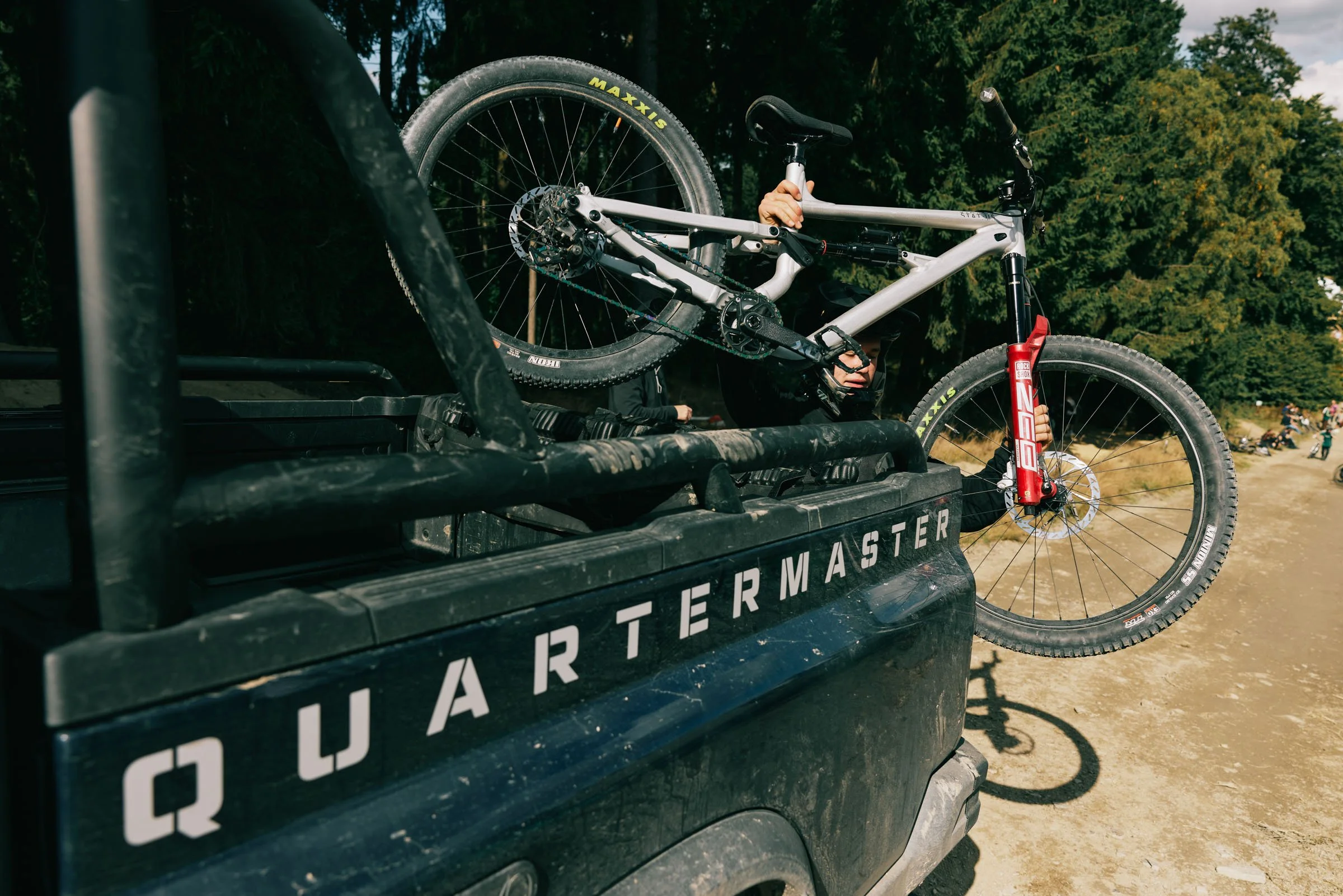 A mountain bike loaded in the bed of a black Jeep Quarantine vehicle with the Quarantine logo, in a dirt outdoor setting with trees in the background.