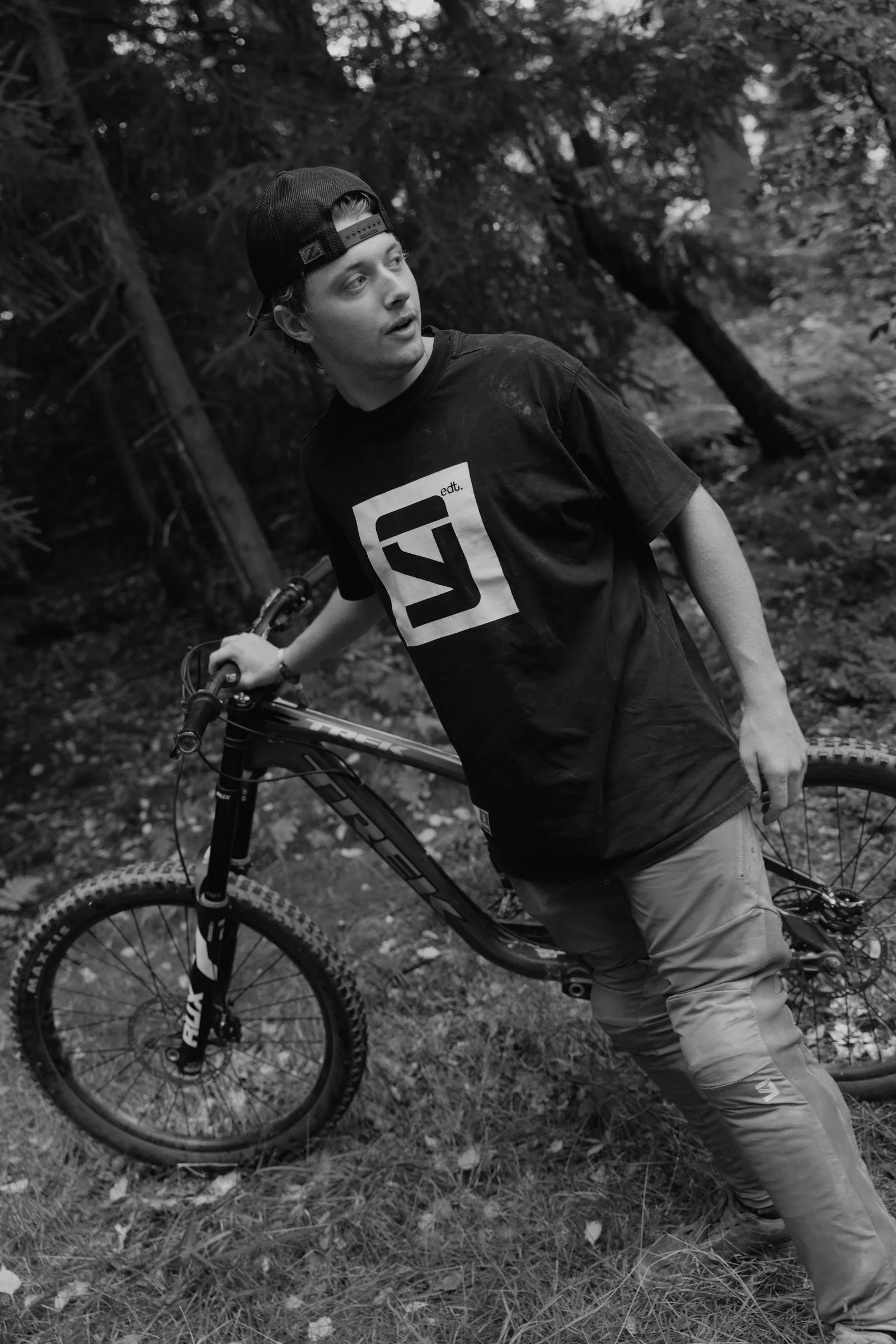Christian Arehart. A young man with a cap and a black T-shirt standing in a forest, holding a mountain bike with one hand, looking to the side.