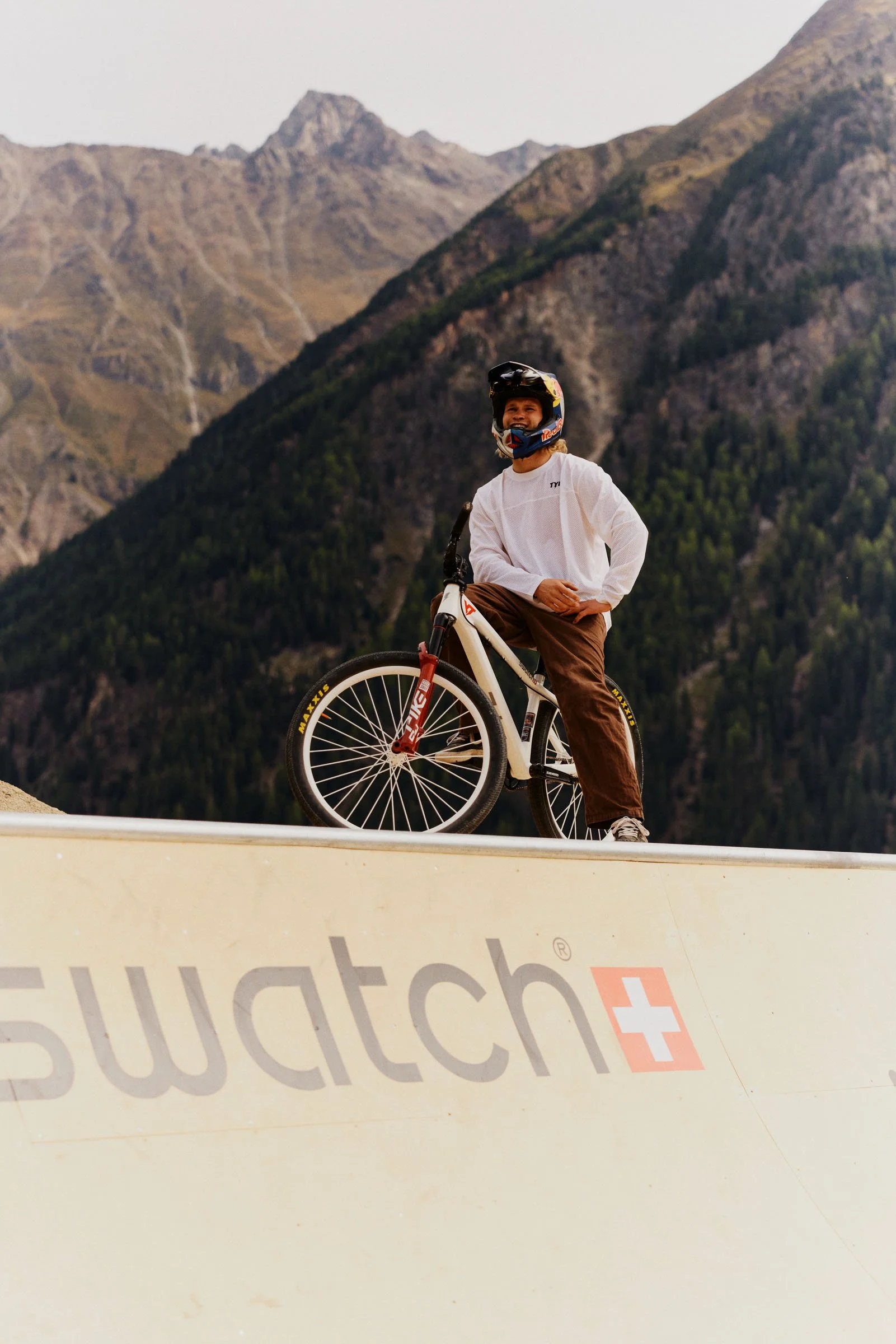 Erik Fedko. A young person wearing a helmet and a white shirt, standing with a mountain bike on a skate park ramp with mountains in the background.