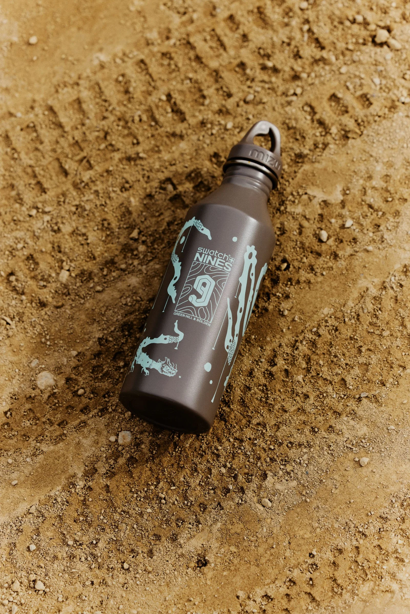 A grey metal water bottle with artistic snake and paint splatter designs, lying on sandy ground.