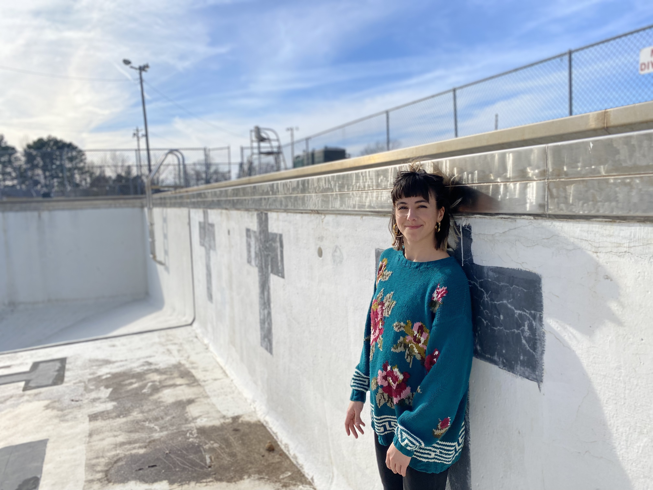 Sam Farquharson, wearing an oversized floral sweater stands in an empty pool, smiling toward the camera, squinting in the sunshine.