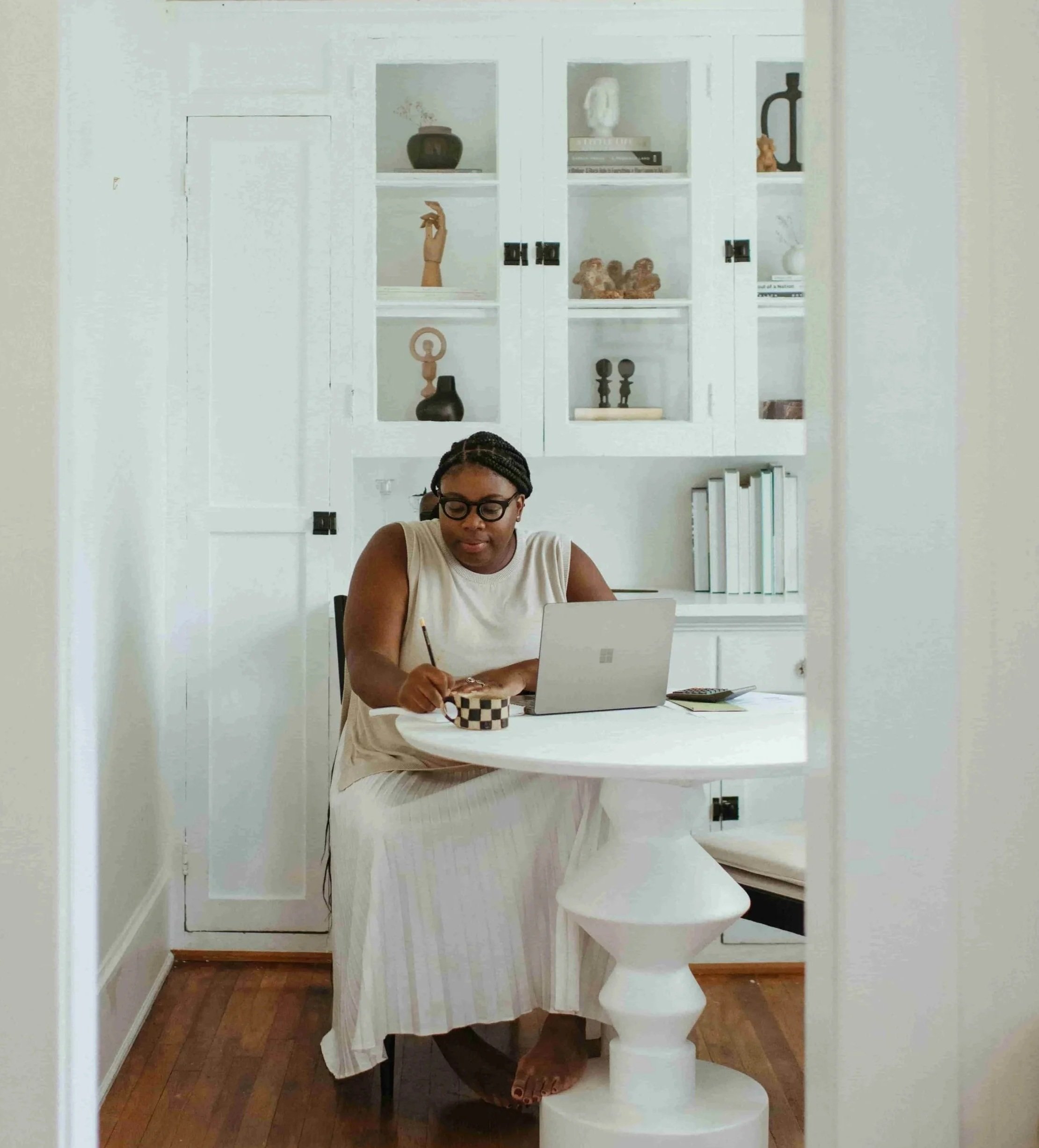 A woman with glasses writing in a checkered container at a white round table, with a laptop and a calculator, in a room with white walls and shelves displaying decorative items, books, and art pieces.