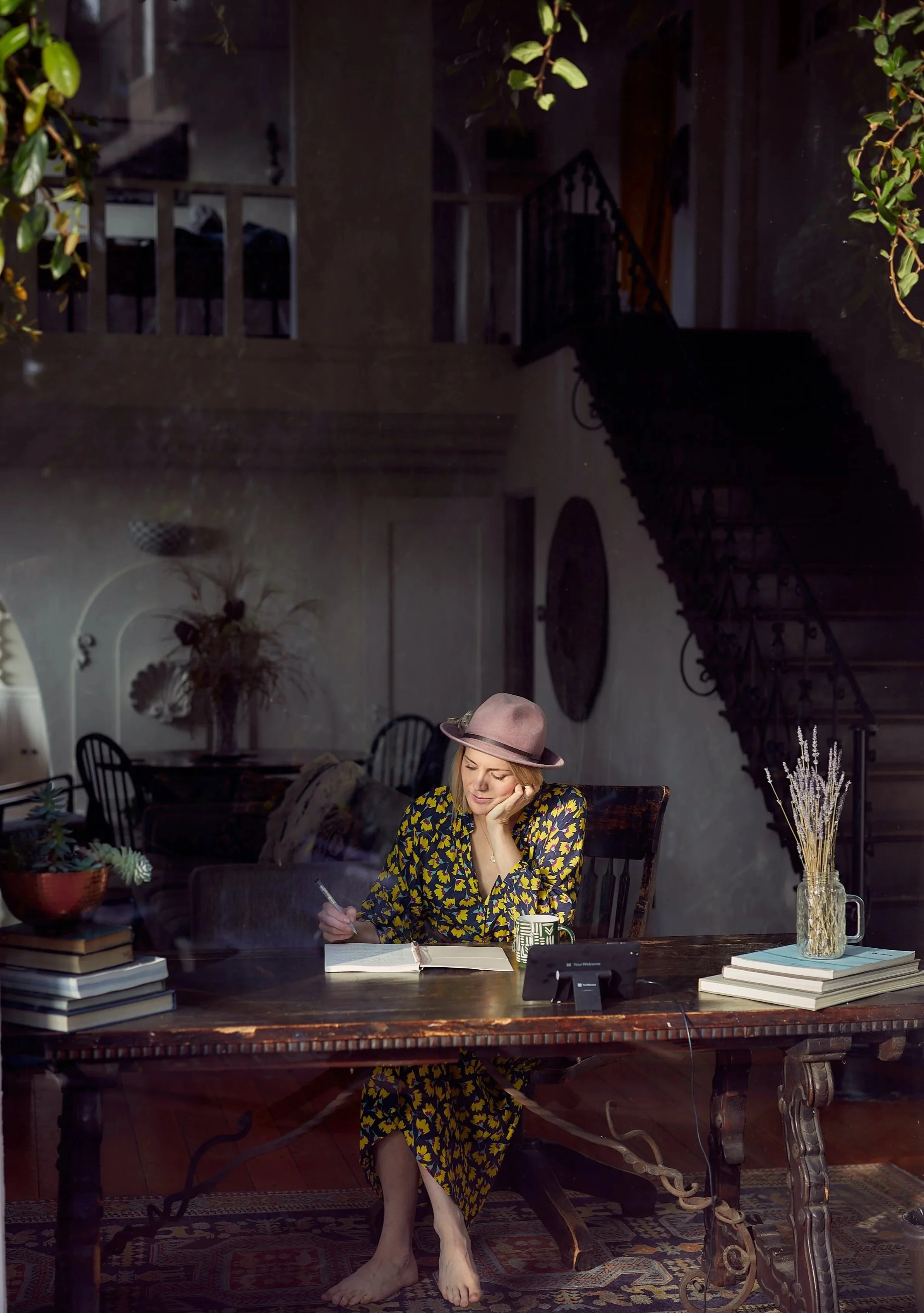 woman in a green and yellow dress in a hat sitting at a desk alone working. She's surrounded by beautiful architecture, books, plants, and antique furniture.