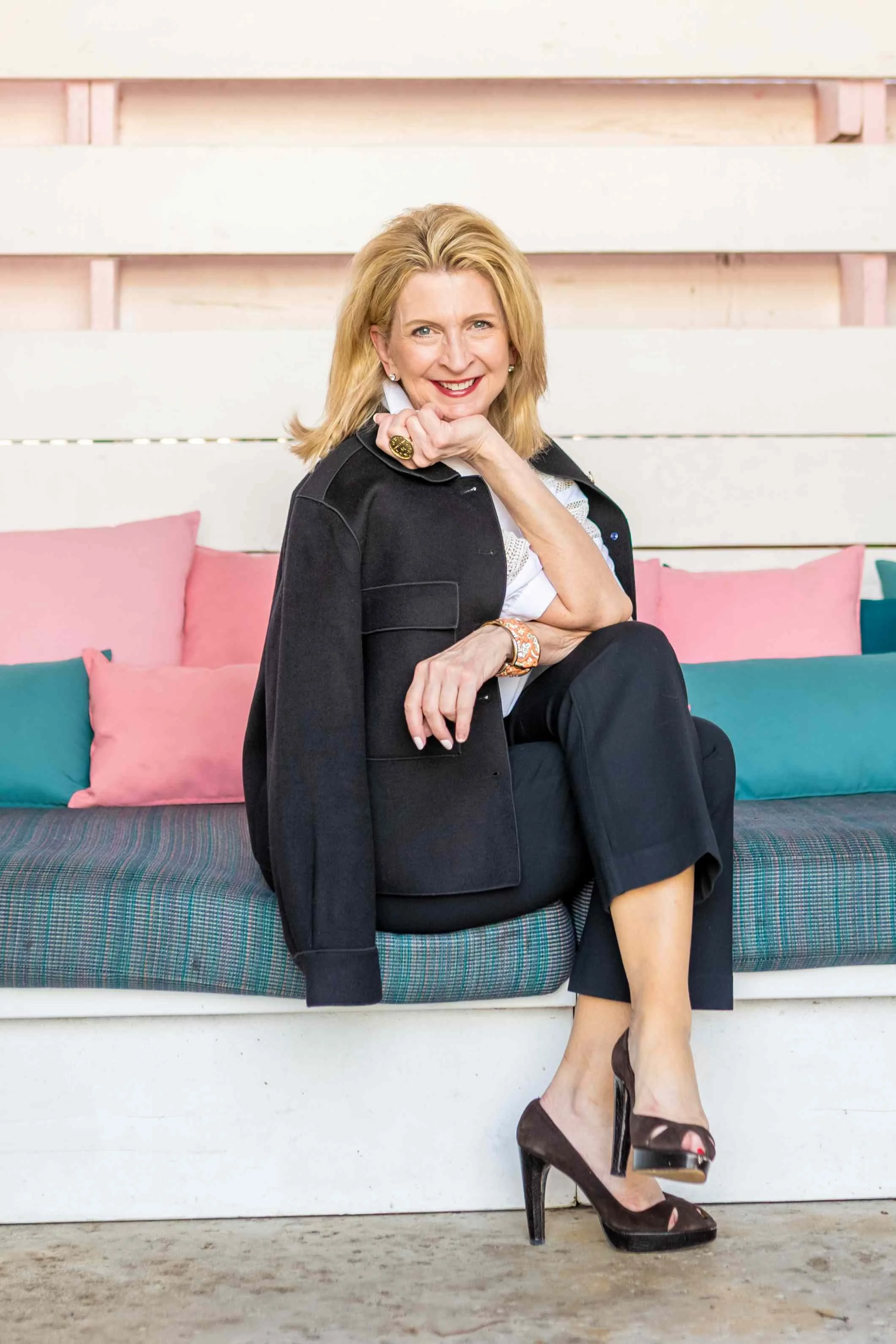 Kimberly in high heels and a black suit, sitting outside on a pink bench with cushions and pillows.