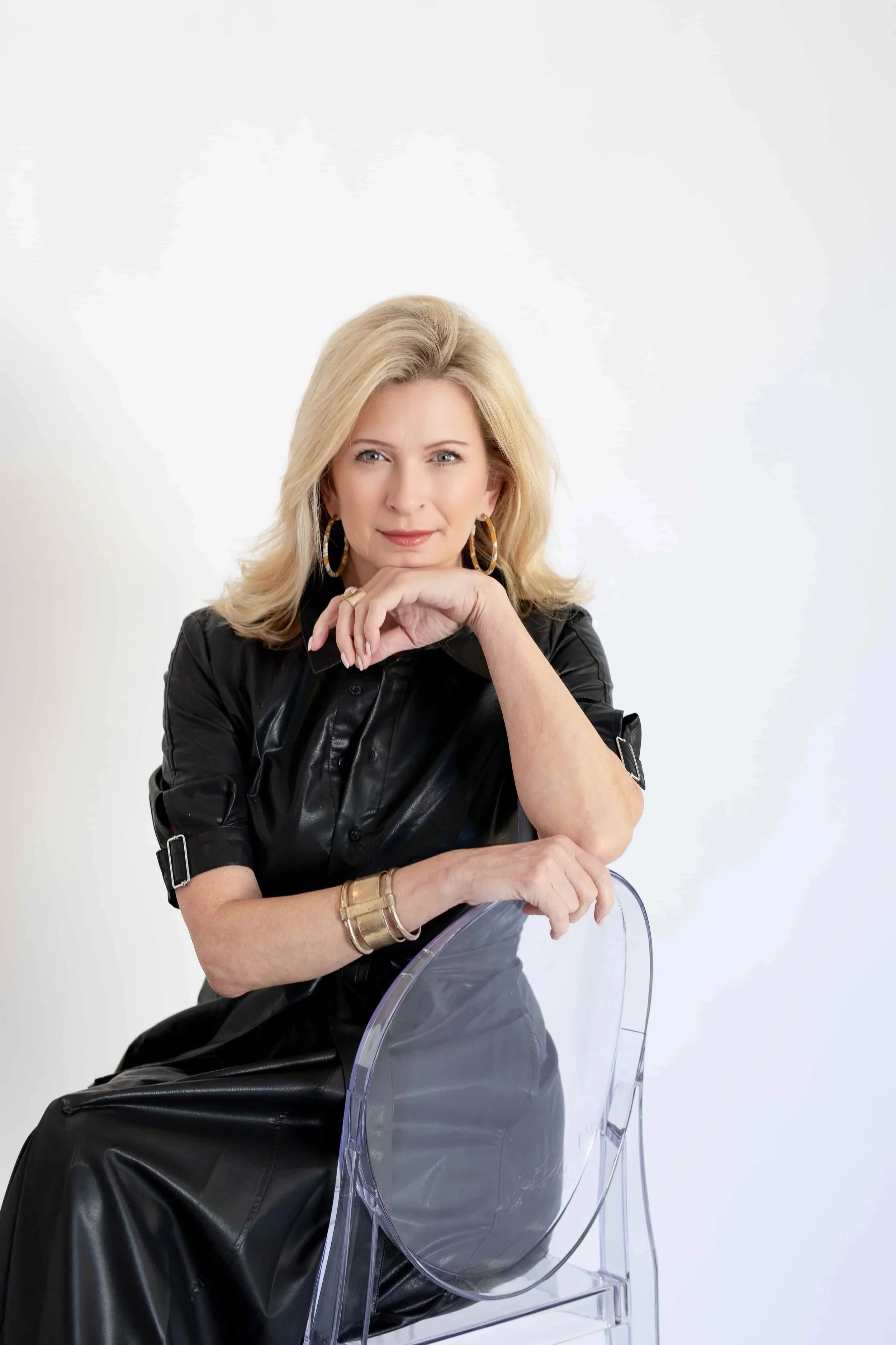 Kimberly Sundt  wearing a black leather dress, gold hoop earrings, and gold bracelets, sitting on a transparent chair against a plain white background, with her hand resting under her chin.