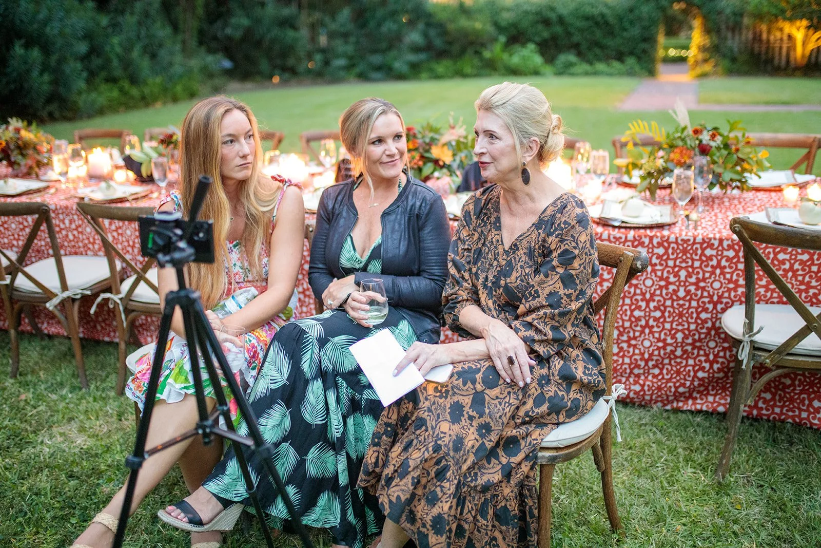 Three women at a retreat sitting at a decorated outdoor event table engaged in conversation, with a camera on a tripod in the foreground.