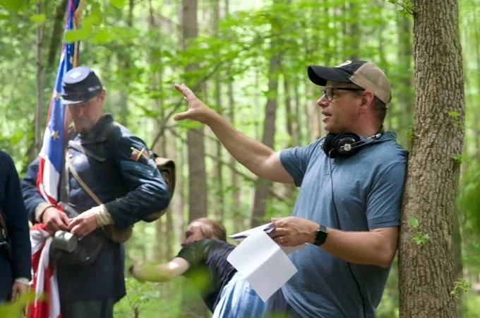 A man wearing a cap, glasses, and headphones is speaking and gesturing with his right hand in a wooded area, while another person dressed in historical clothing holding a flag stands nearby.
