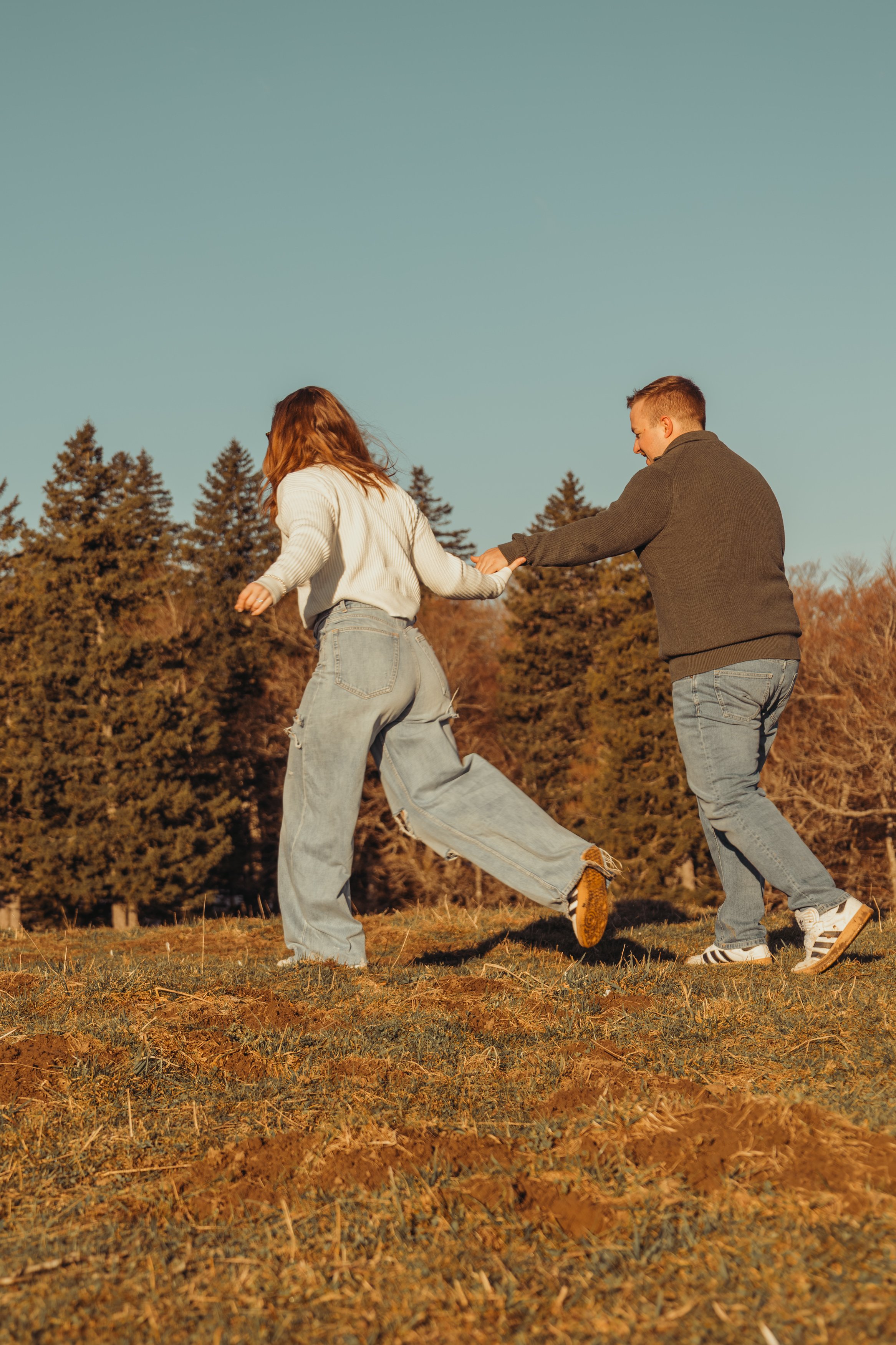 Deux personnes (un homme et une femme) jouent et s'amusent dans un champ d'automne, tenant la main. L'homme porte un pull gris et un jean, la femme porte un pull blanc et un jean déchiré. Le ciel est clair et un groupe d'arbres en arrière-plan.
