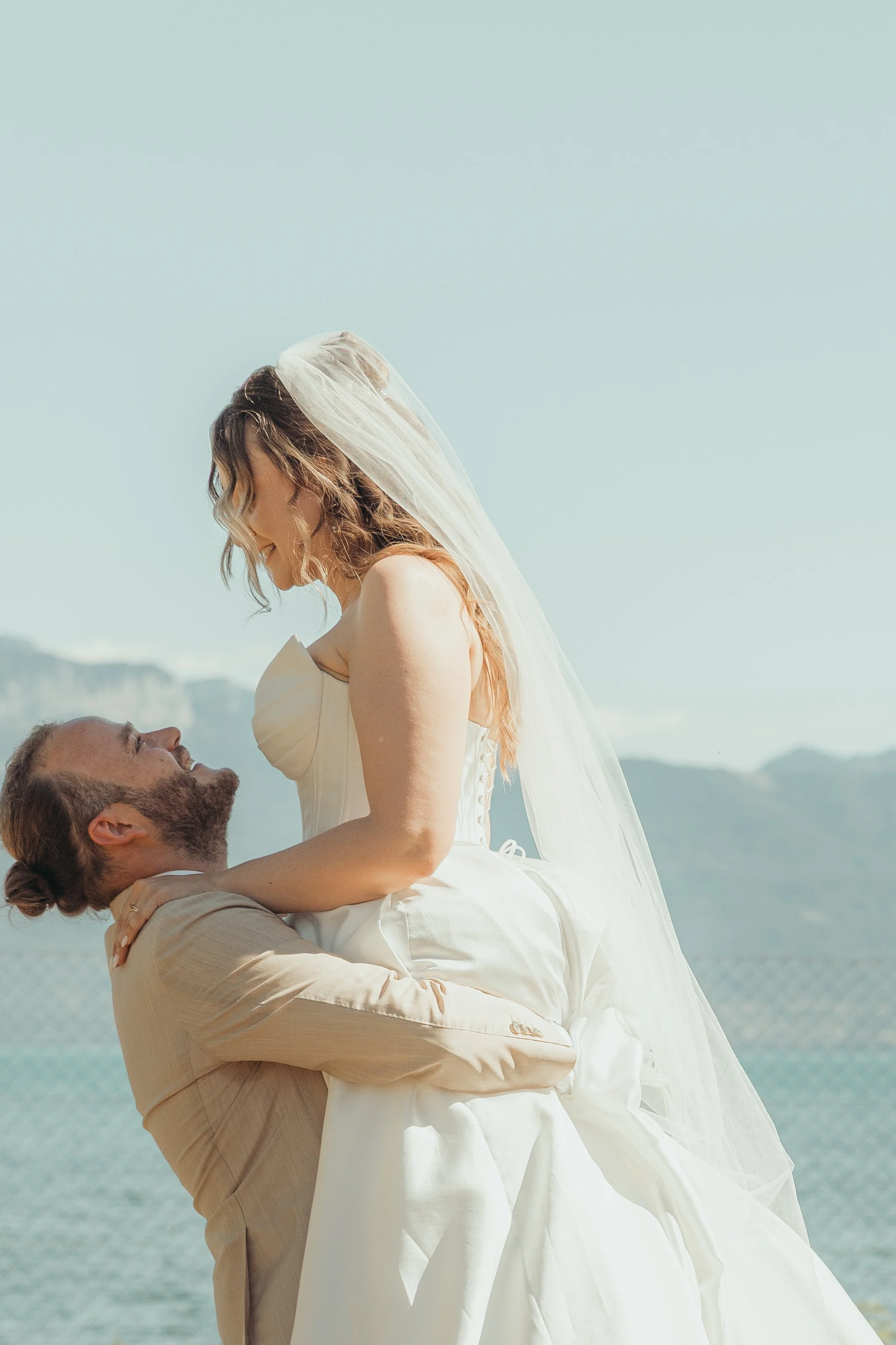 Un couple marié en pleine célébration, avec la femme portant une robe de mariée blanche et un voile, et l'homme portant un costume beige, devant un paysage naturel avec la mer et des montagnes en arrière-plan.