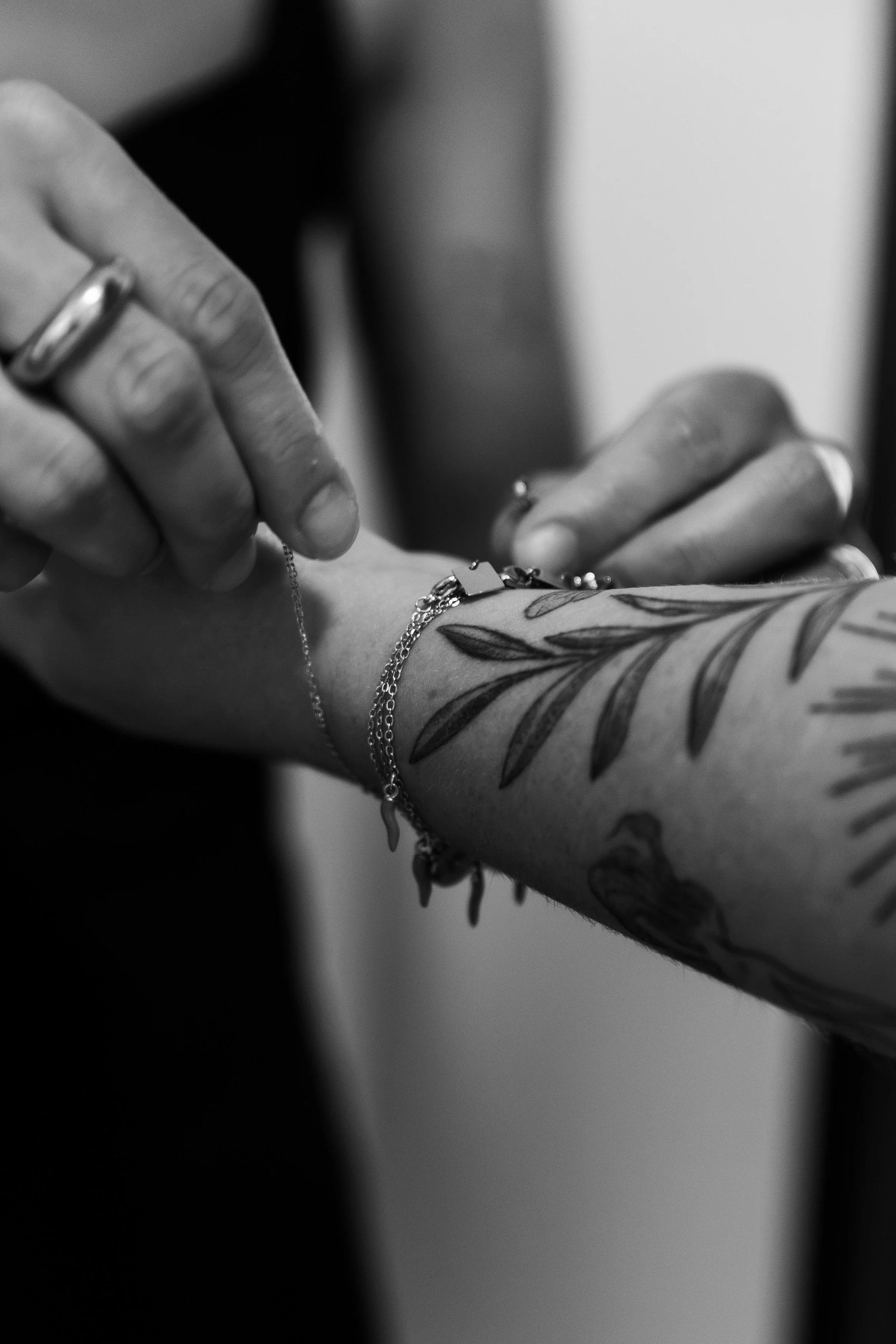 Une personne ajuste une chaîne autour du poignet, visible une tatouage de feuilles, des bagues et des bracelets en argent.