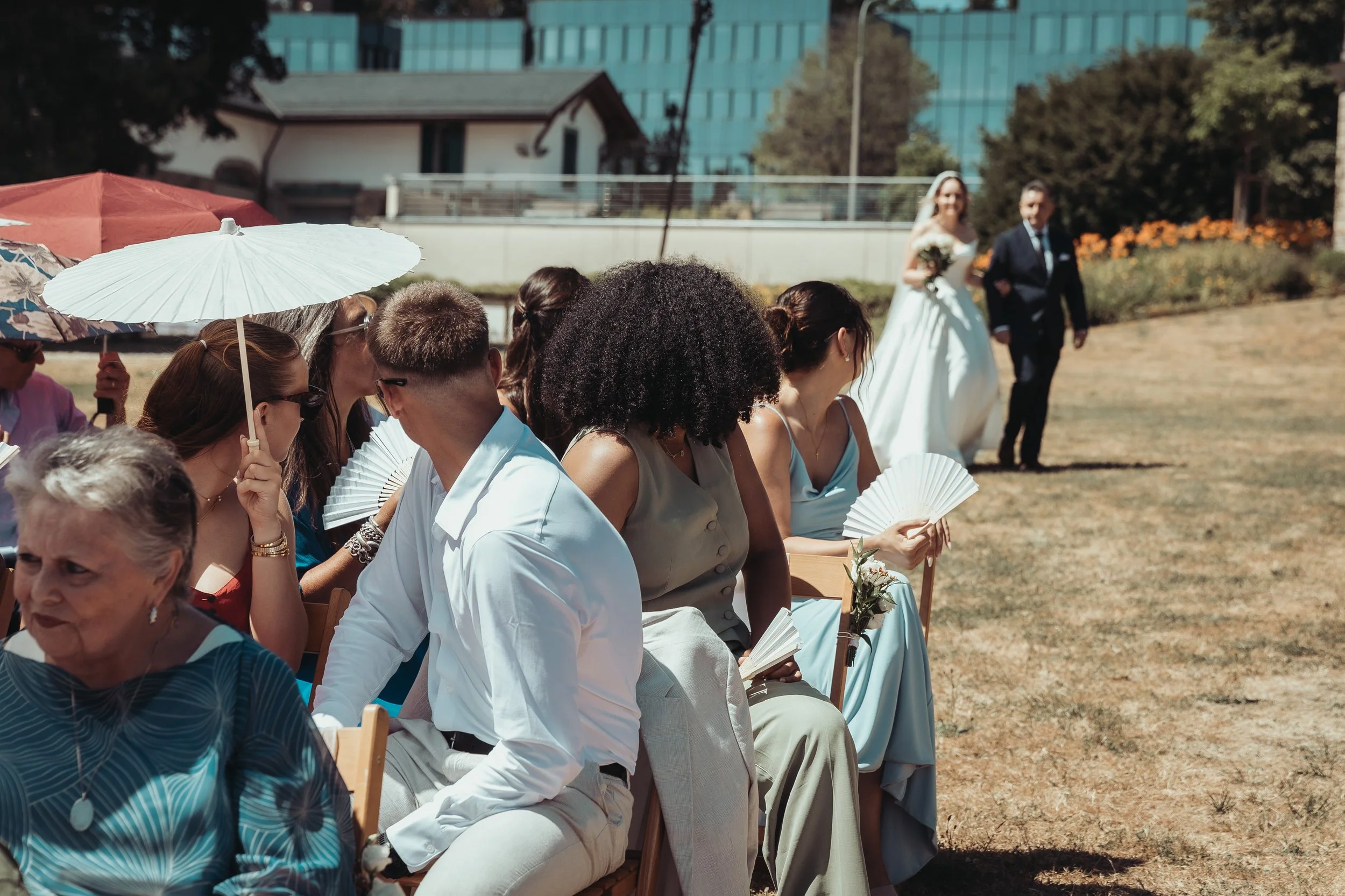 Cérémonie de mariage en extérieur avec des invités assis, certains tenant des parapluies, fond d'une ville moderne. La mariée en robe blanche et le marié en costume marchent vers l'avant.