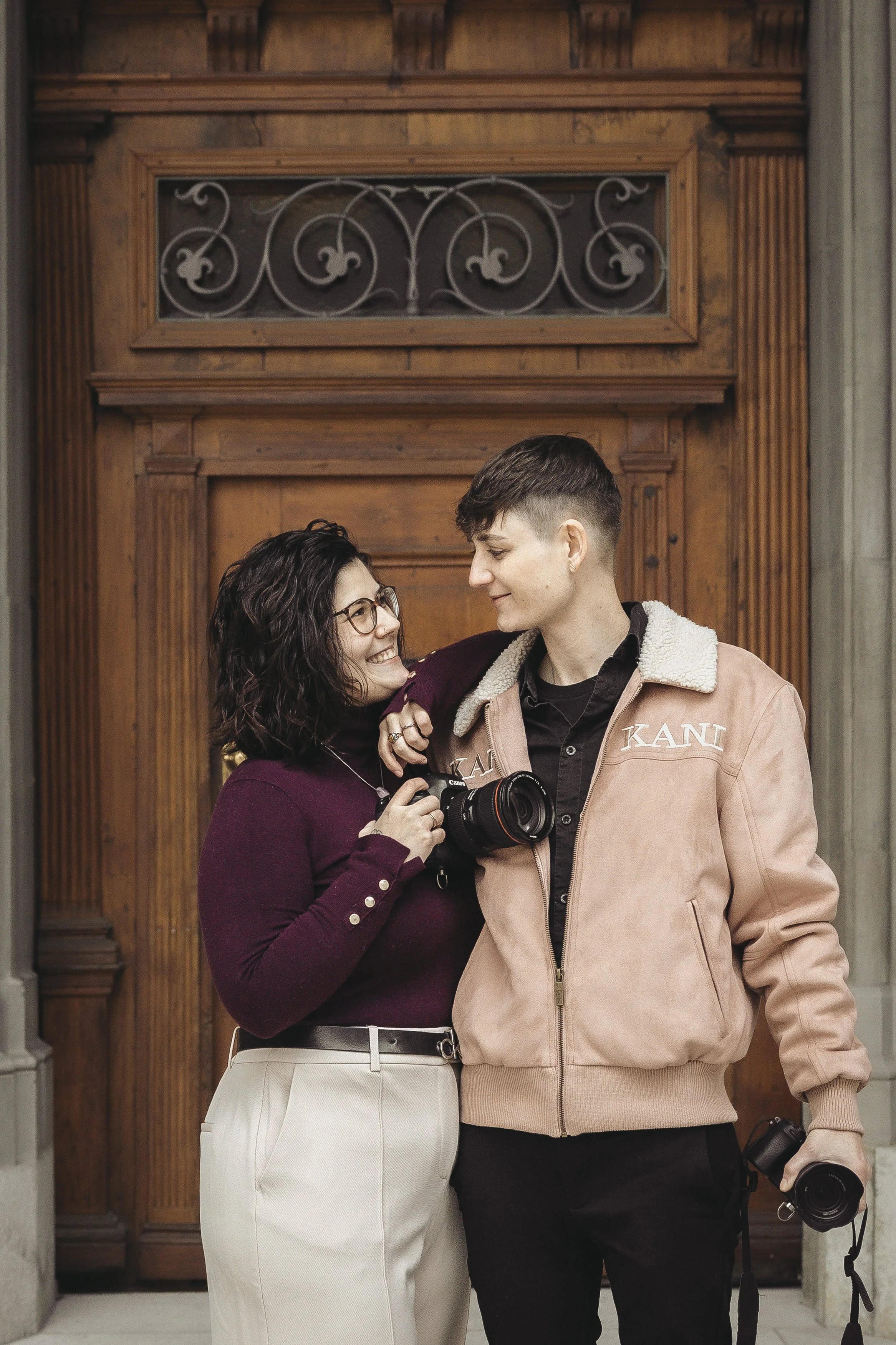 Deux jeunes adultes avec des appareils photo se regardant et souriant devant une porte en bois.