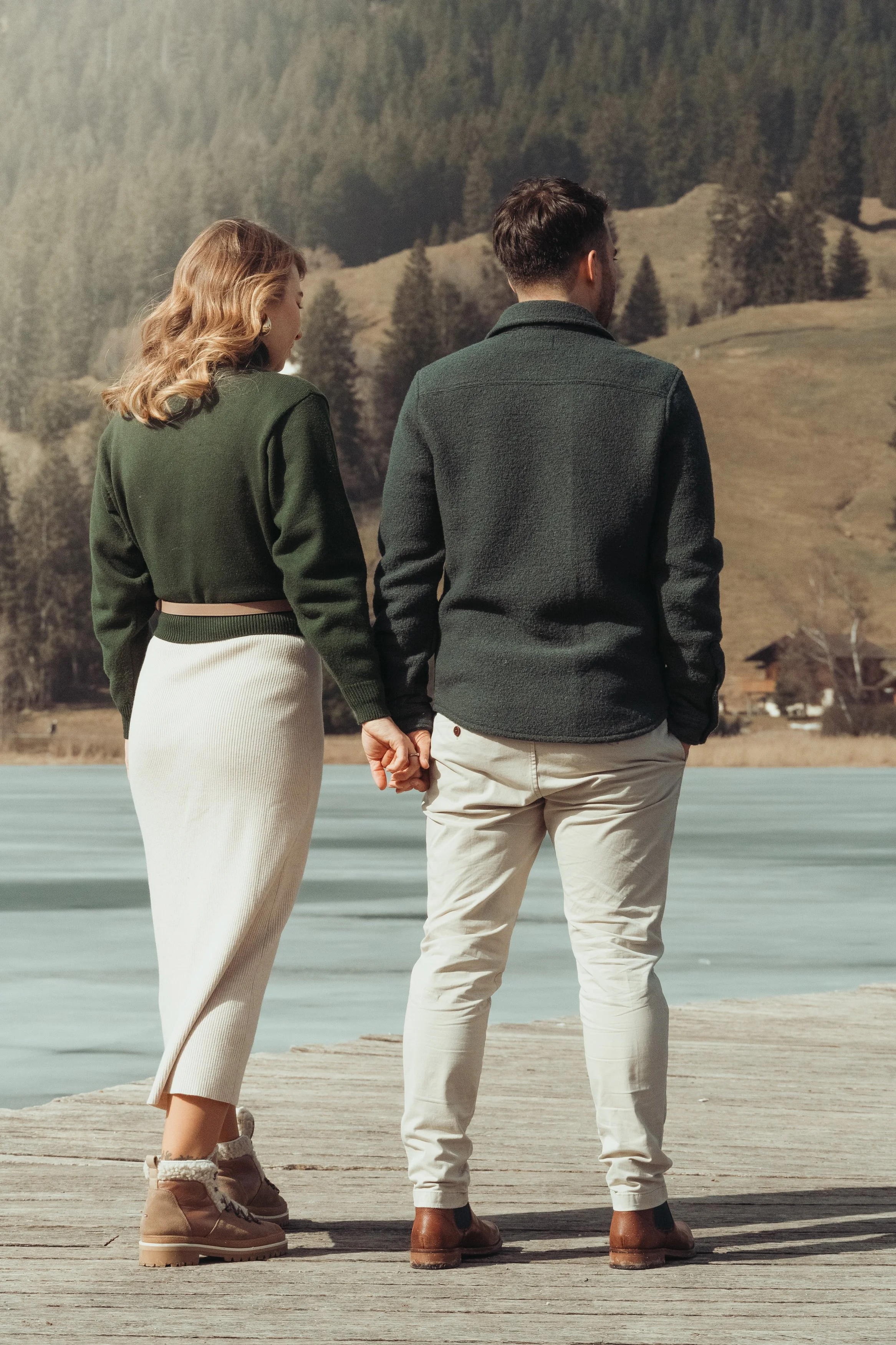 Un couple se tient la main sur un quai en bois près d'un lac ou d'une rivière, avec des montagnes et des arbres en arrière-plan, tenant la main et regardant dans la même direction.