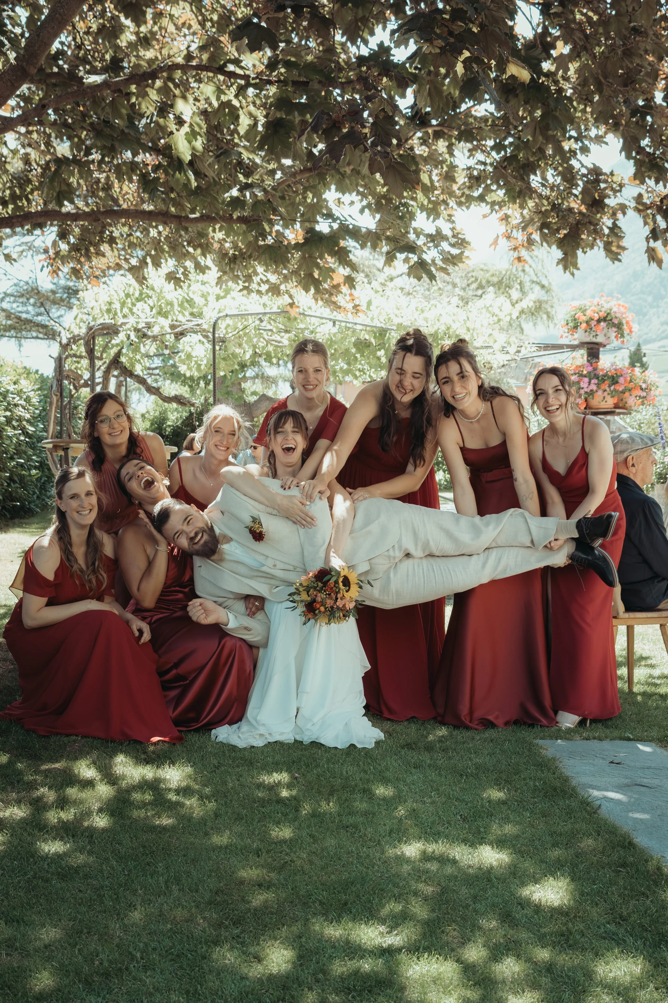 Un groupe de personnes en vêtements de mariage et de demoiselle d'honneur posant en extérieur sous un arbre, avec des fleurs en arrière-plan.