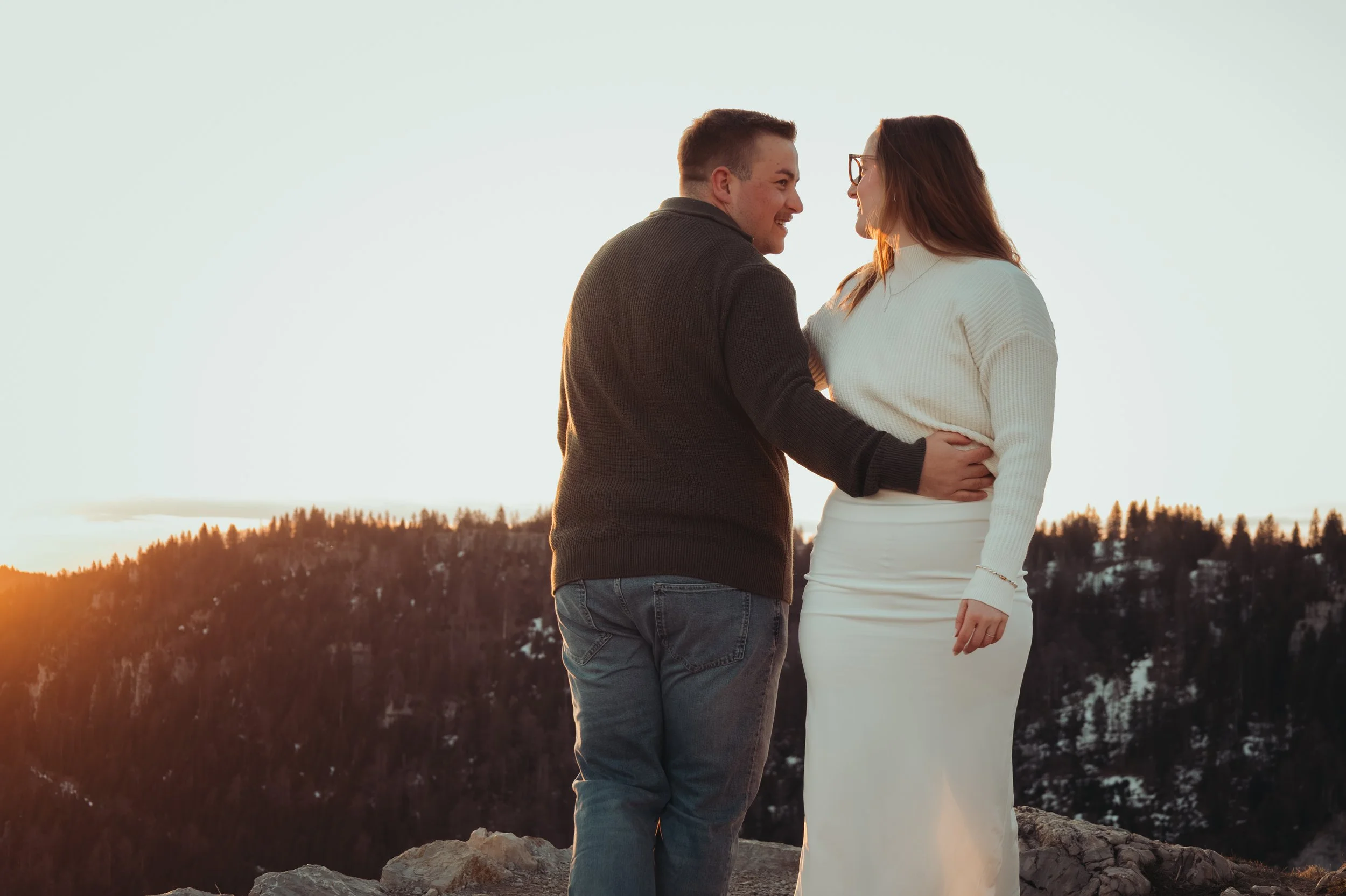 Un couple regardant avec affection l'un l'autre lors d'un coucher de soleil dans un paysage montagneux, lui en pull gris et elle en robe blanche, partageant un moment romantique.