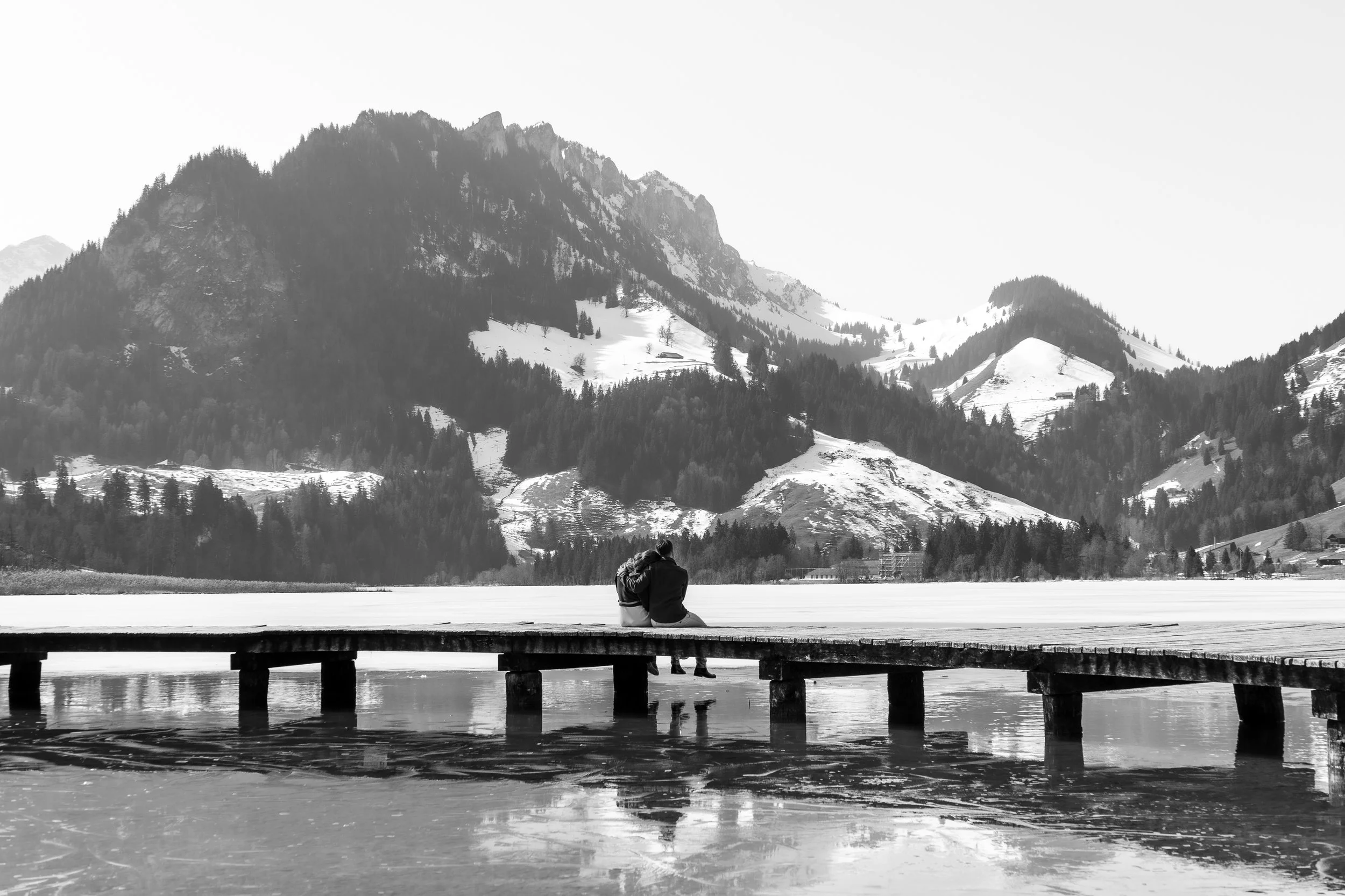 Deux personnes assises sur un pont en bois au bord d'un lac, regardant des montagnes enneigées en arrière-plan, en noir et blanc.