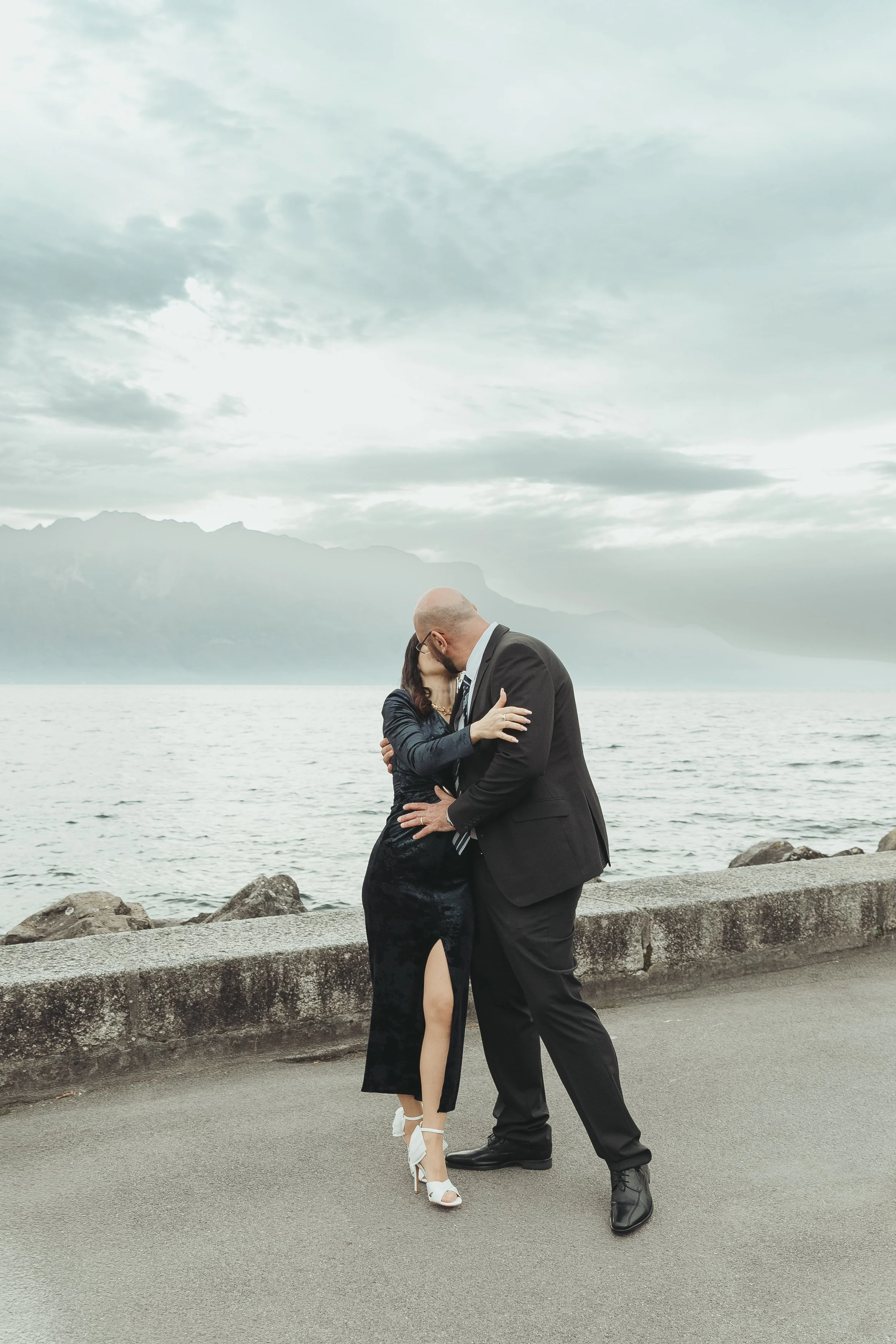 Des mariés s'embrassant près d'une mer, avec des montagnes en arrière-plan et un ciel nuageux lors d'un reportage mariage avec Marie Monod photographe d'émotions, duo de photographes queer en Suisse.