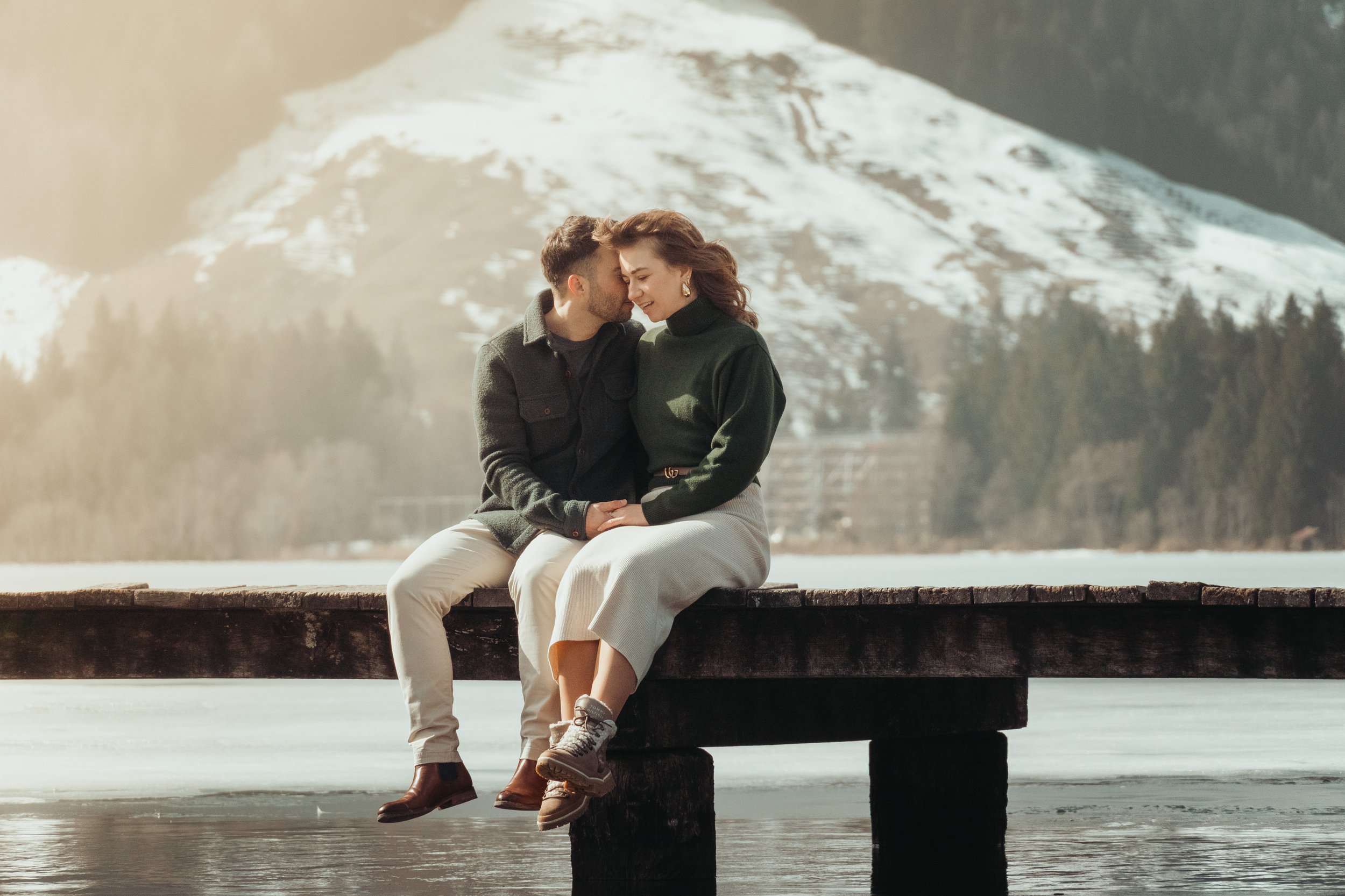 Un couple assis sur un quai en bois au bord d'un lac, le visage presque touché, partageant un moment intime avec une montagne enneigée en arrière-plan.