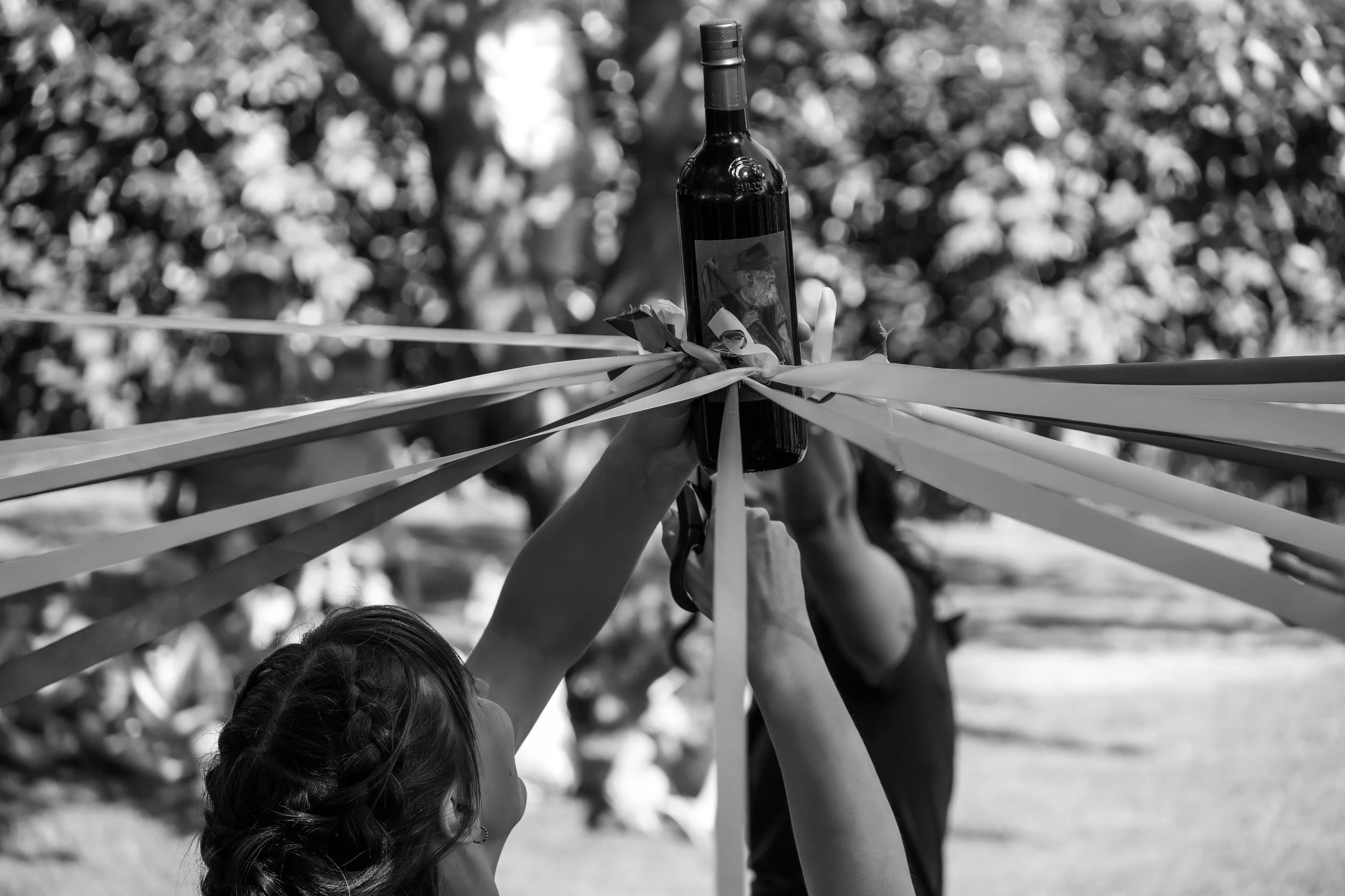 Deux personnes festives, dont une avec des cheveux bruns en tresse, tiennent une bouteille de vin en l'air lors d'une célébration en plein air. Des rubans décoratifs entourent la bouteille.
