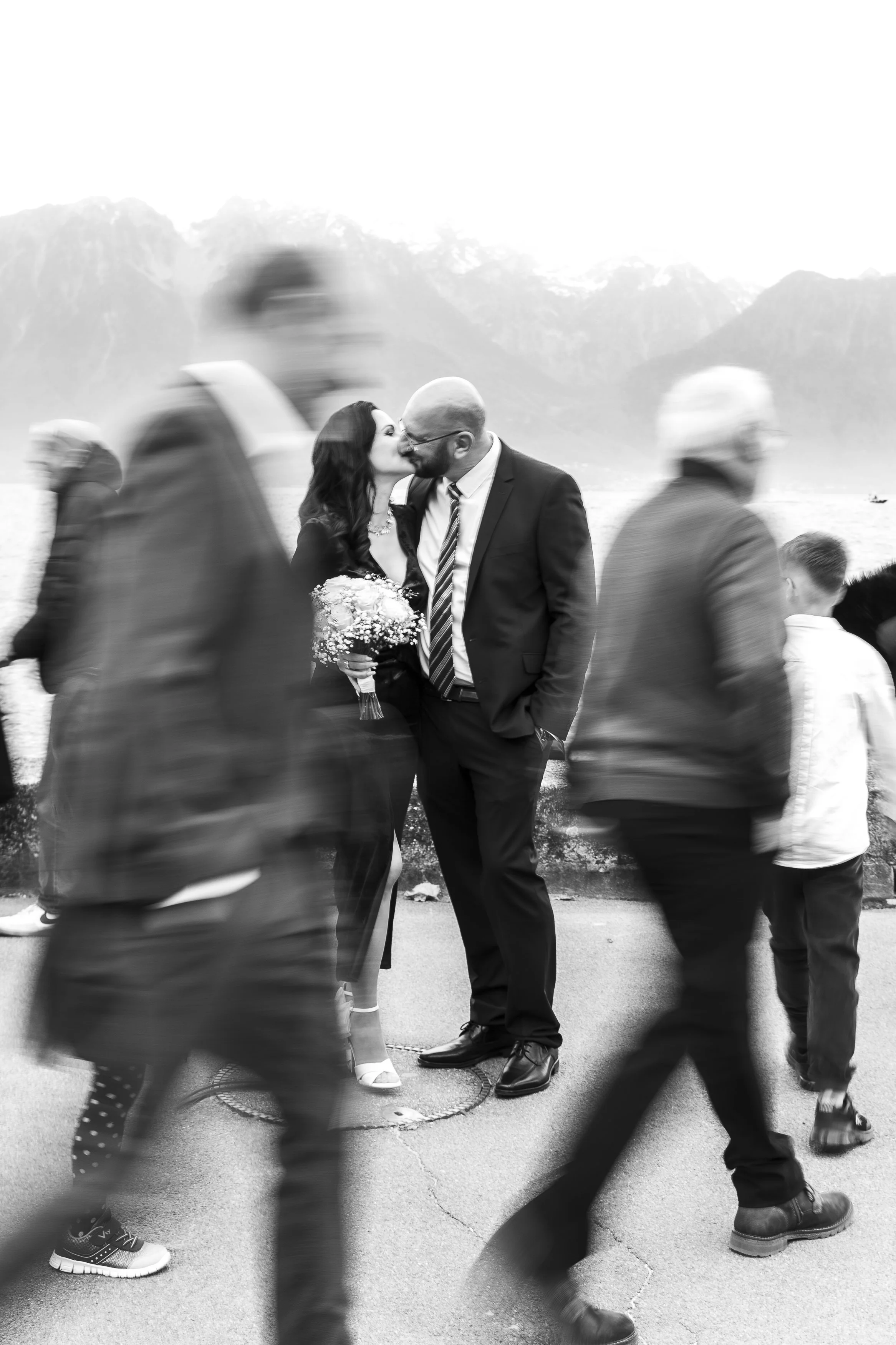 Deux mariés s'embrassent lors d'une cérémonie de mariage en plein air avec un fond de montagnes, entourées d'autres personnes floues qui passent lors d'un reportage mariage avec Marie Monod photographe d'émotions, duo de photographes queer en Suisse.