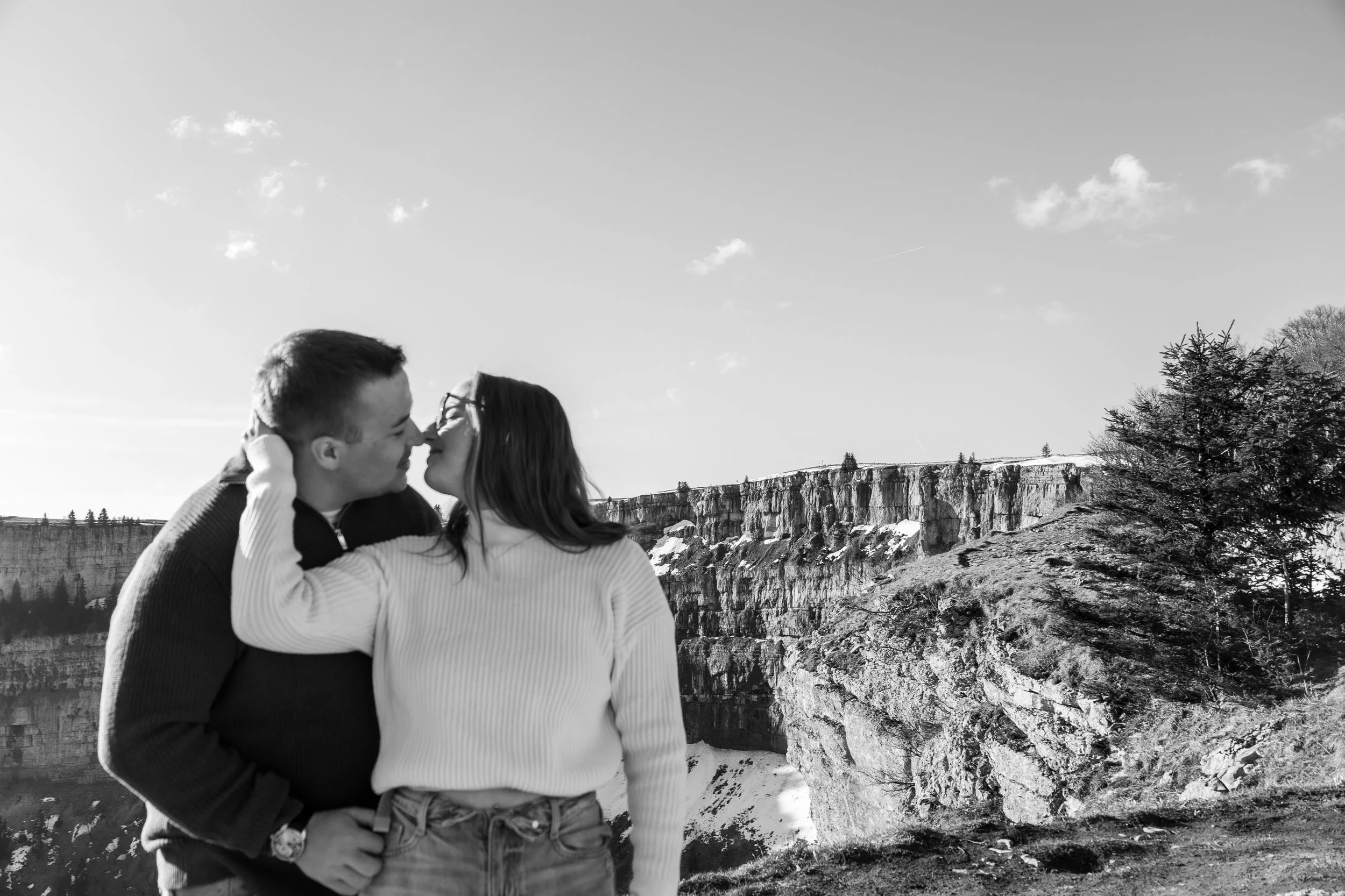Un couple s'embrasse dans un paysage montagneux avec des falaises et des arbres, en noir et blanc.