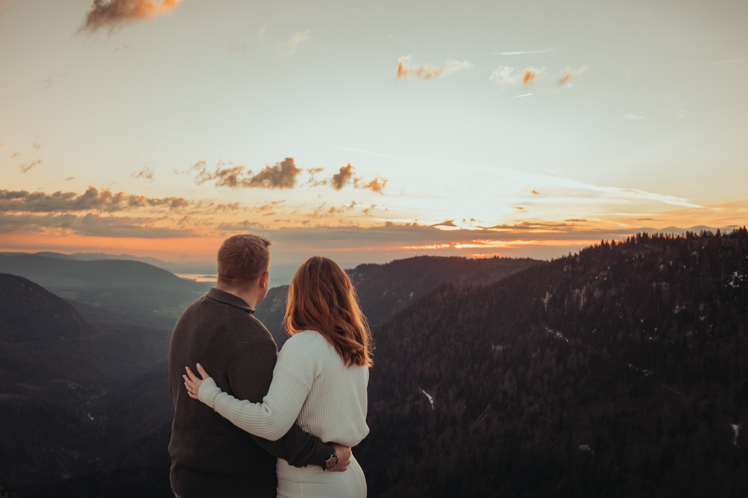 Un couple regarde un coucher de soleil à l'horizon, surplombant une vallée montagneuse, pendant que le ciel est parsemé de nuages.