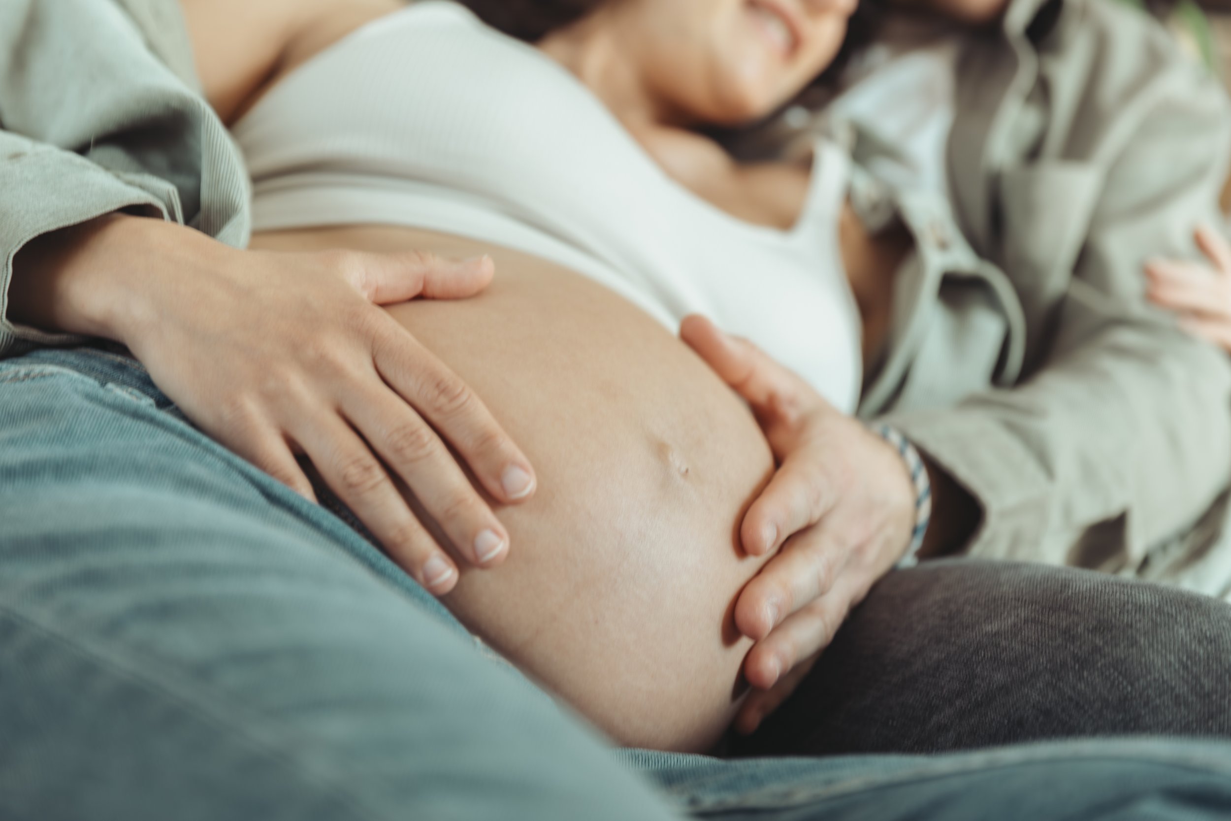 Une femme enceinte est allongée contre son conjoint, les mains sur le ventre lors d'une séance photo grossesse avec Marie Monod photographe d'émotions, duo de photographes queer en Suisse.