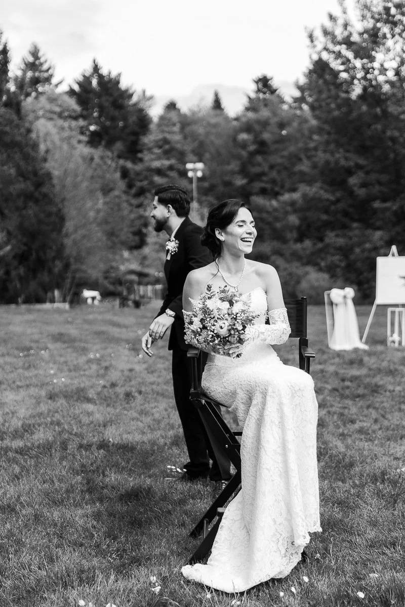 Une photo en noir et blanc d'un mariage en plein air, avec une femme en robe de mariée assise sur une chaise, tenant un bouquet, et un homme en costume marchant derrière elle, tous deux souriants, entourés par la nature.