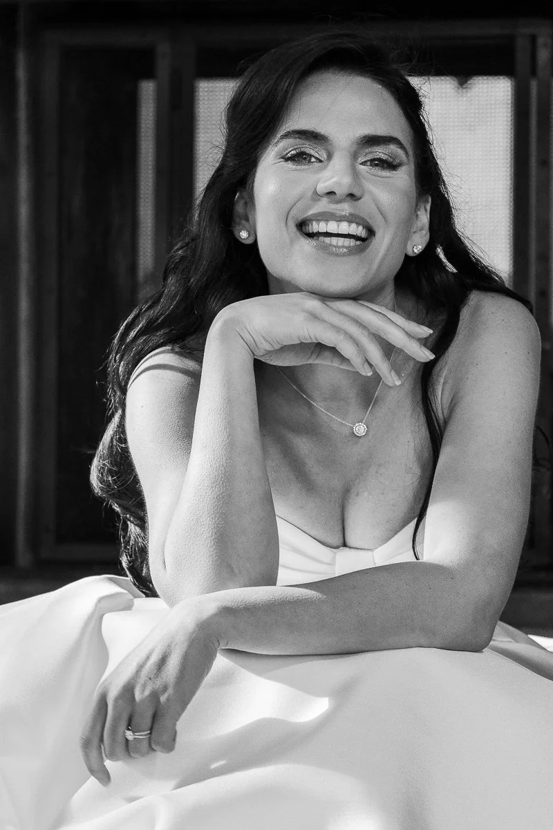 Une femme souriante en robe blanche, posant avec la main sous le menton, dans un style noir et blanc.