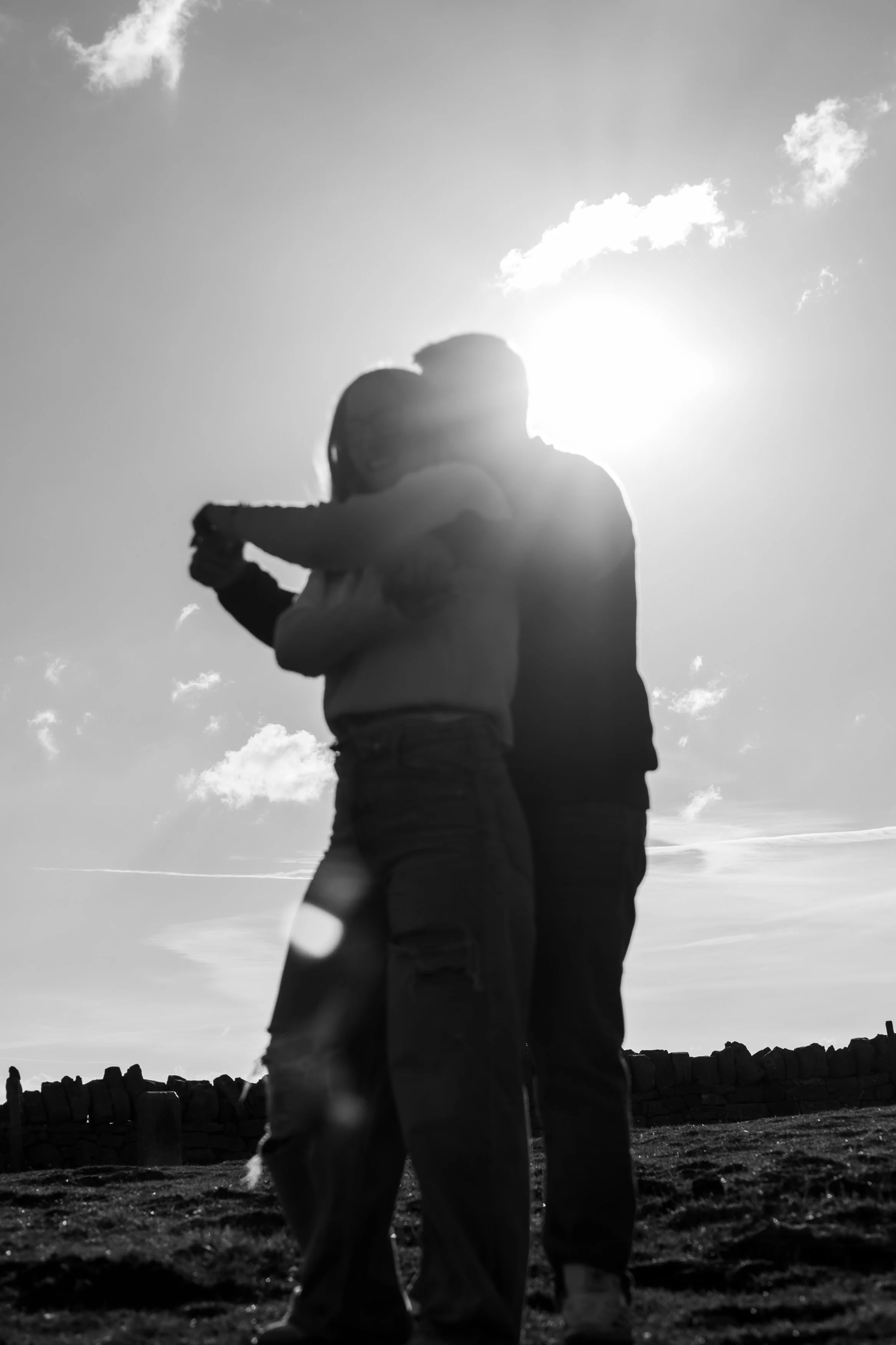 Deux personnes qui s'embrassent ou qui se donnent un câlin, photographiées en silhouette avec le soleil en arrière-plan, dans un décor extérieur avec une mirador ou un mur en pierre.
