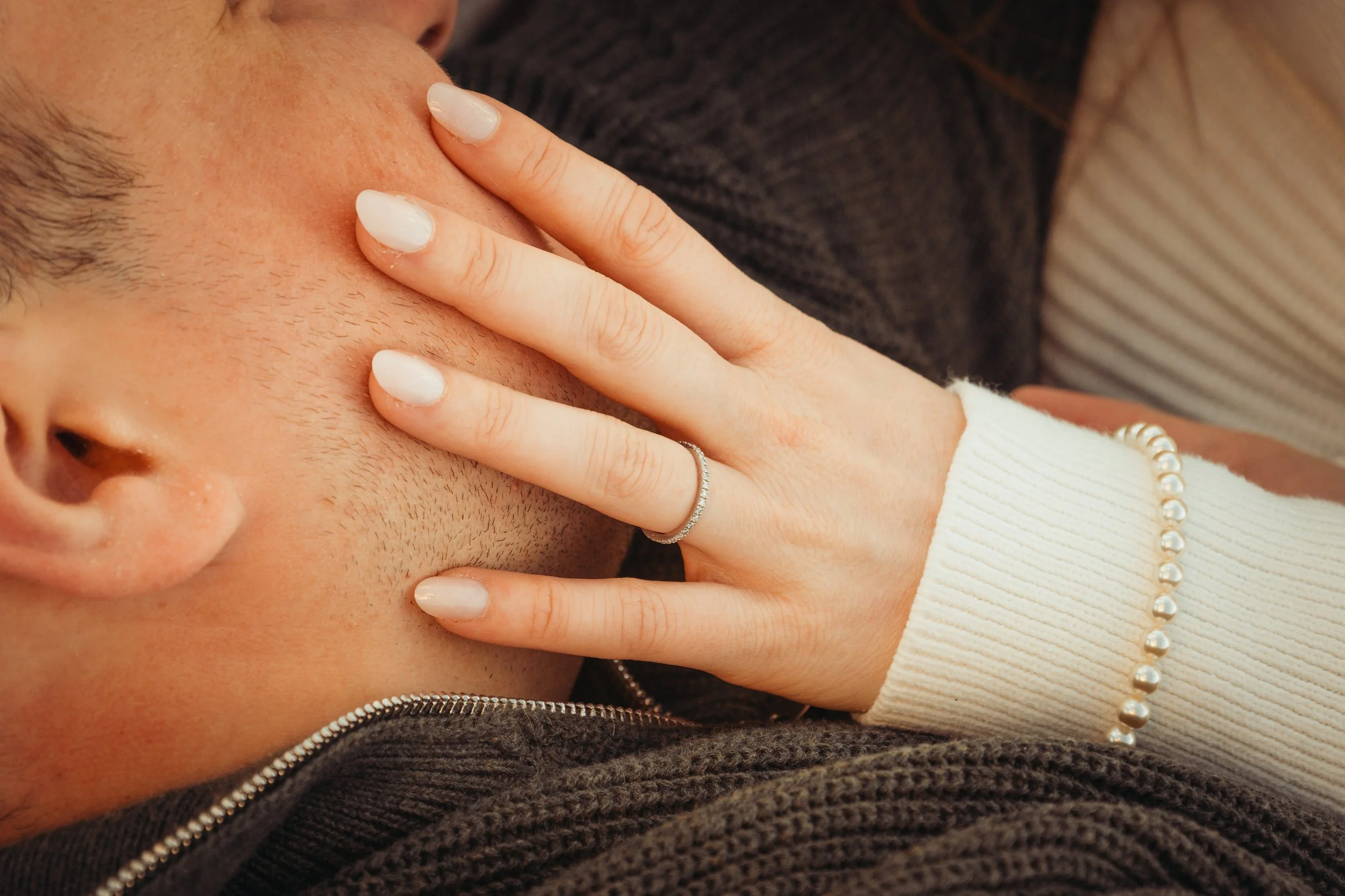 Une personne avec une barbe qui est touchée au visage avec ses doigts. Elle porte un pull blanc avec un bracelet en perles. La personne porte aussi une bague discrète et un haut sombre.