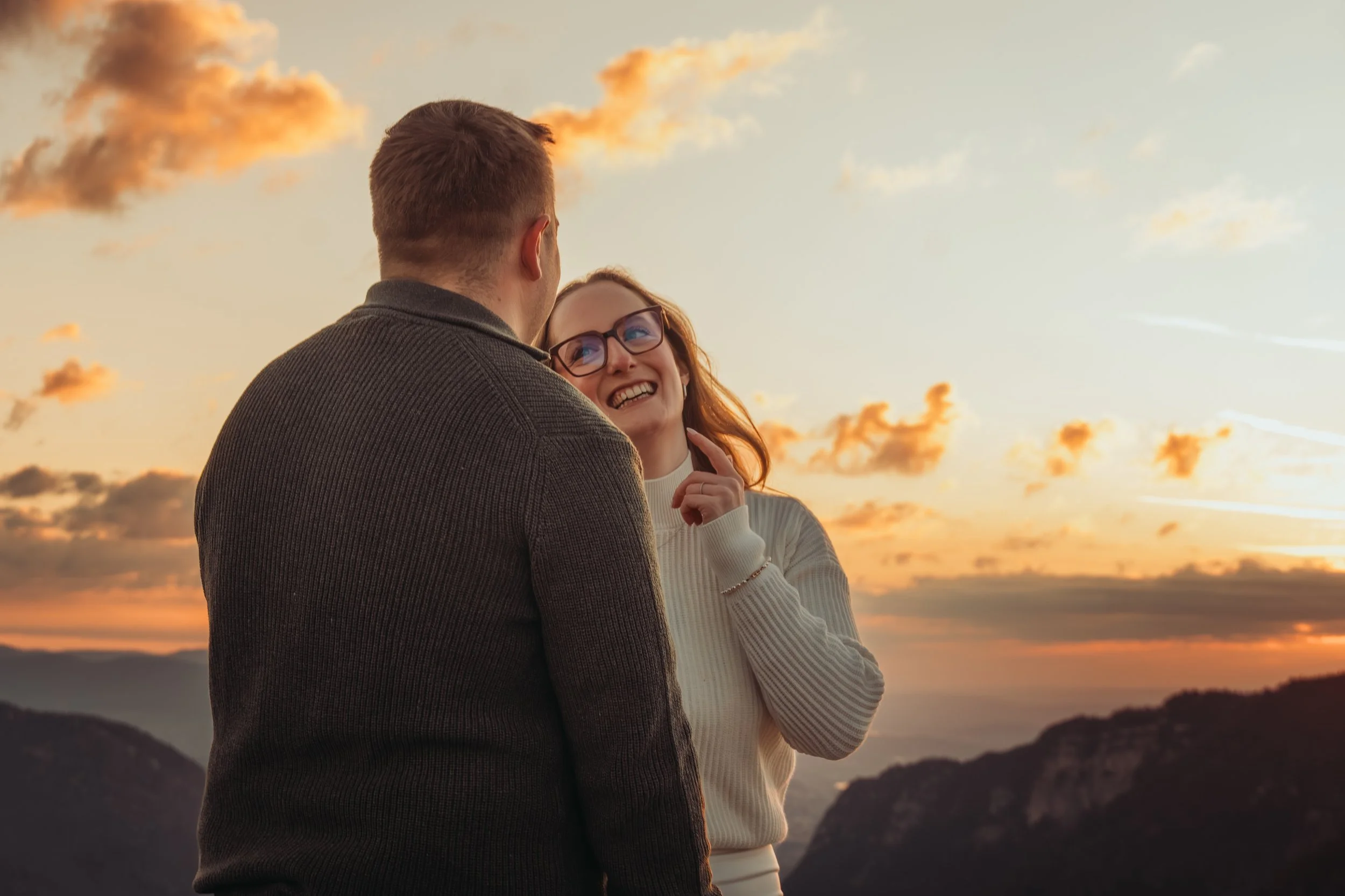 Un couple souriant lors d'un coucher de soleil, avec des montagnes en arrière-plan, partage un moment de bonheur ensemble.