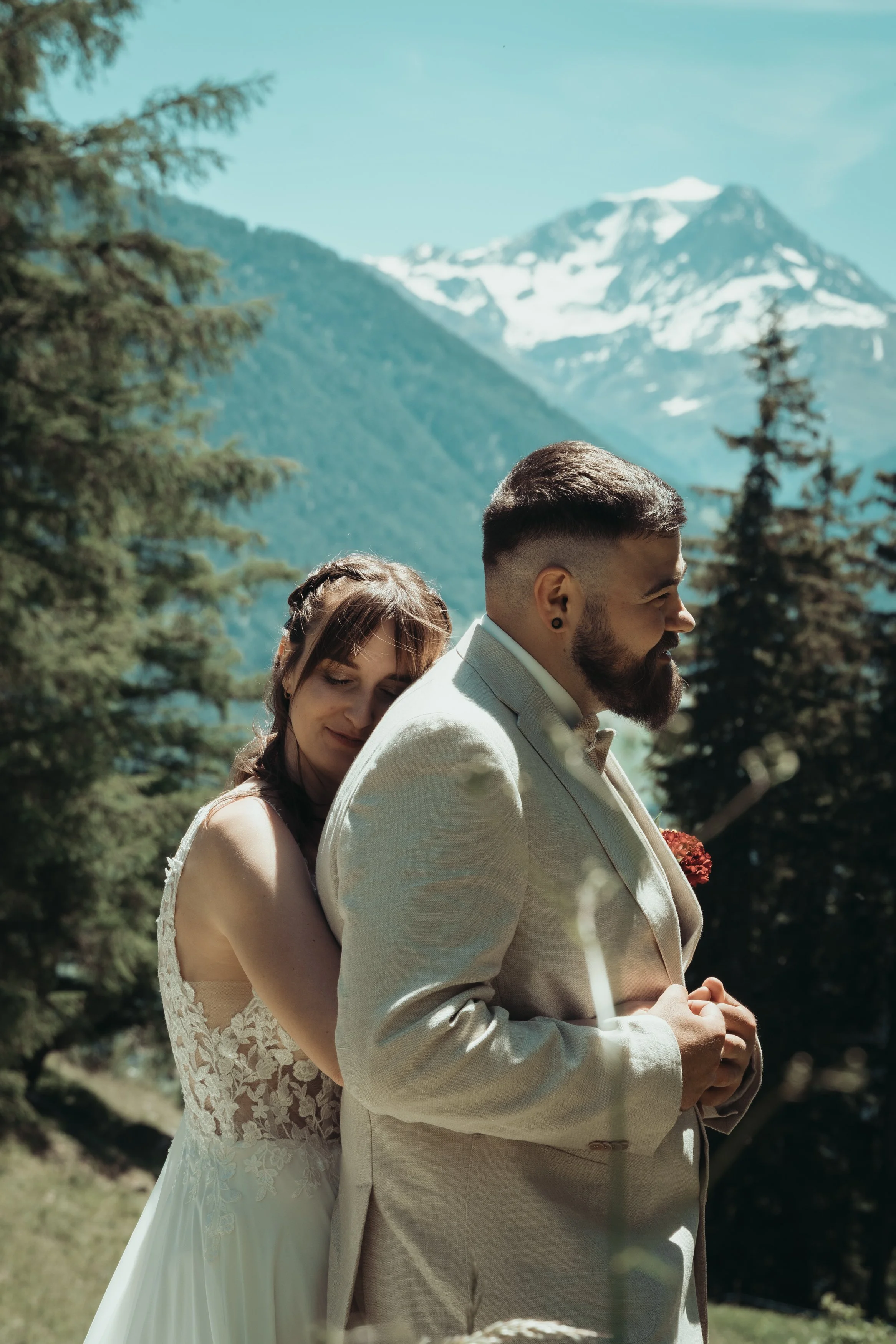 Un couple en habits de mariage, la femme en robe blanche à dentelle embrassant le dos de l'homme en costume beige, dans un paysage de montagne avec des arbres et un sommet enneigé en arrière-plan.