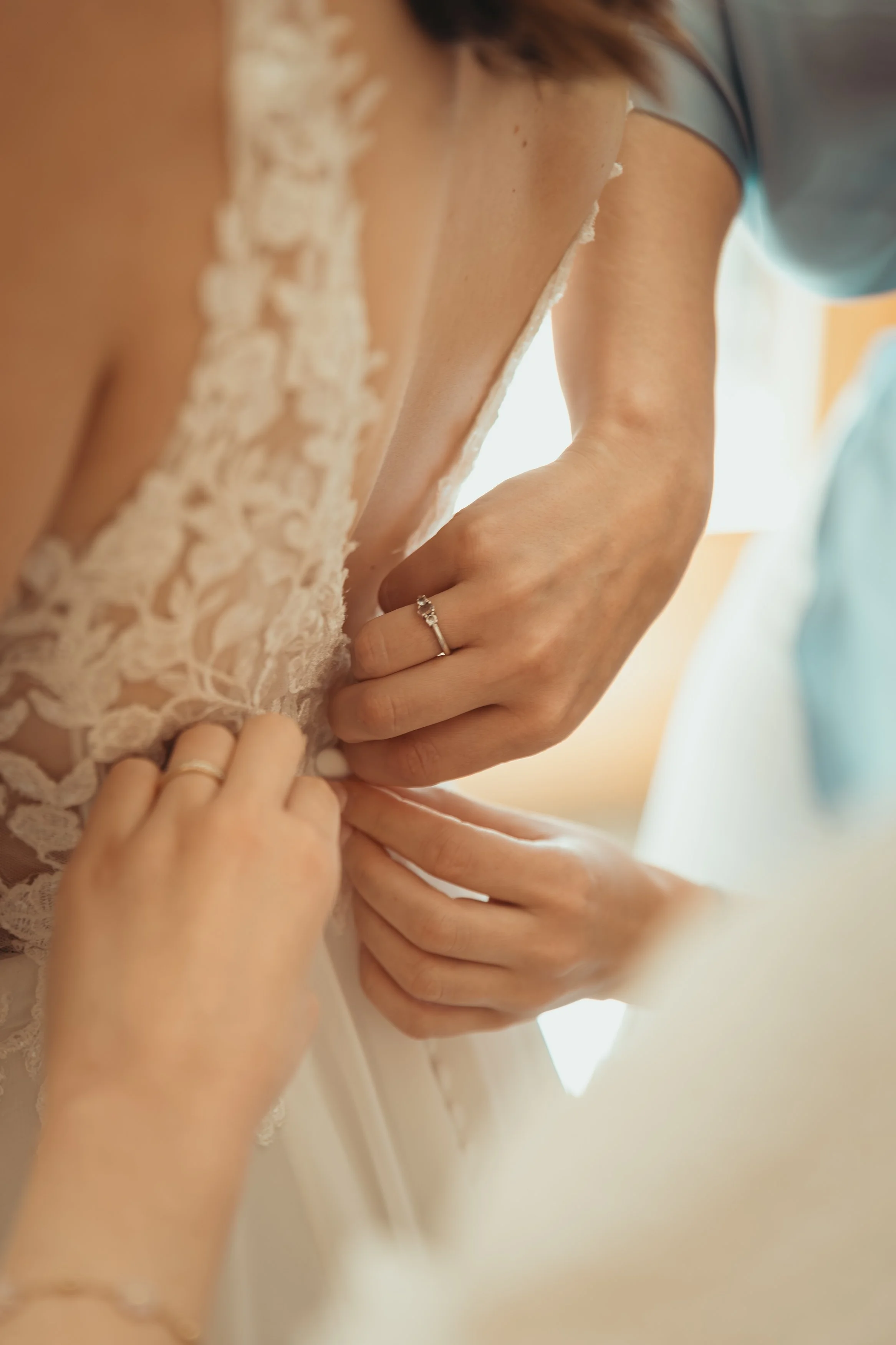Une femme en robe de mariée ajustant son corsage, avec des mains qui l’aident, portant une bague. La scène est intime et délicate, capturant un moment de préparation nuptiale.