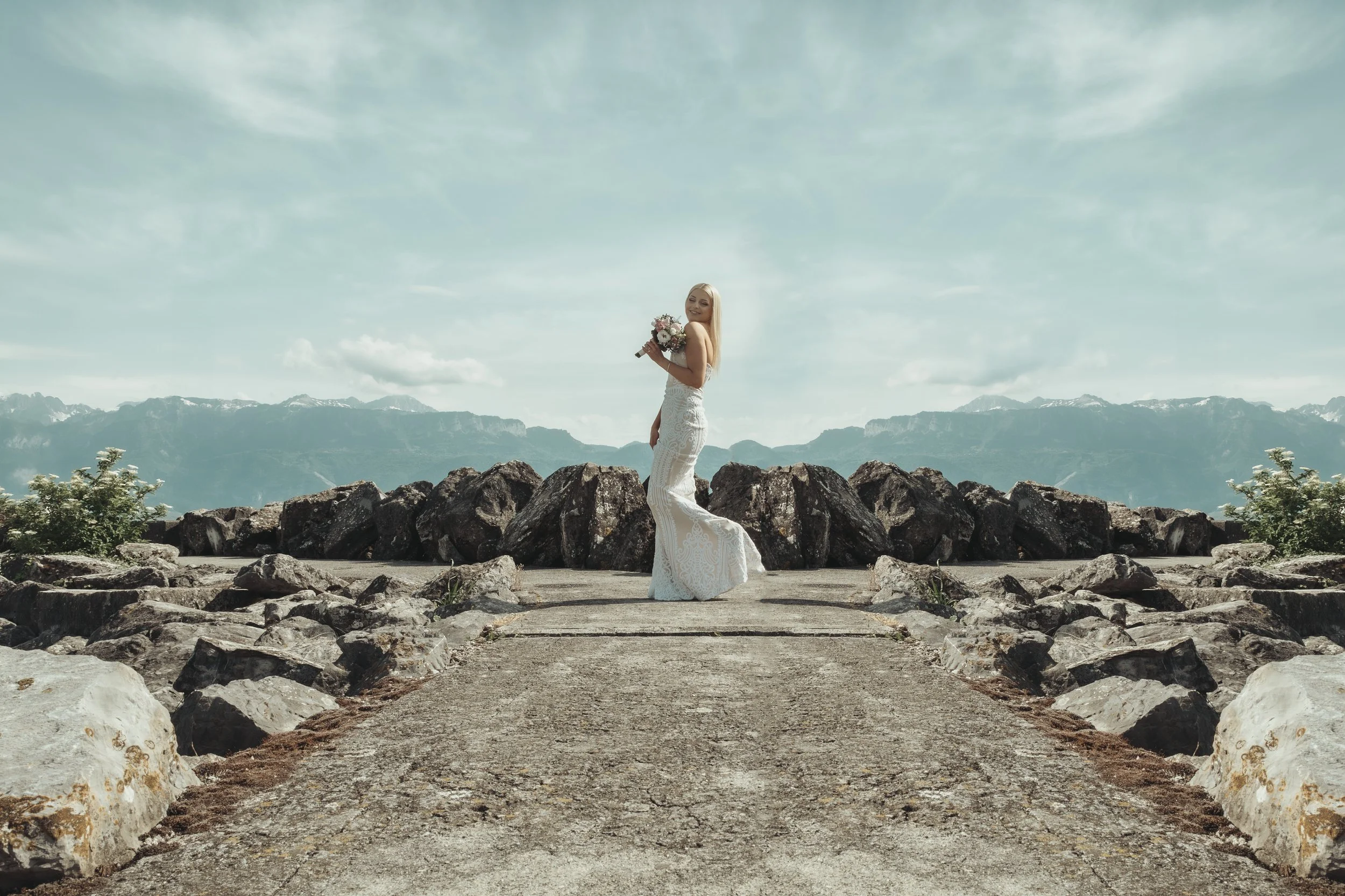 Une femme en robe de mariage tient un bouquet de fleurs dans un décor montagnard avec des rochers, un chemin en pierre et des montagnes en arrière-plan, sous un ciel partiellement nuageux.