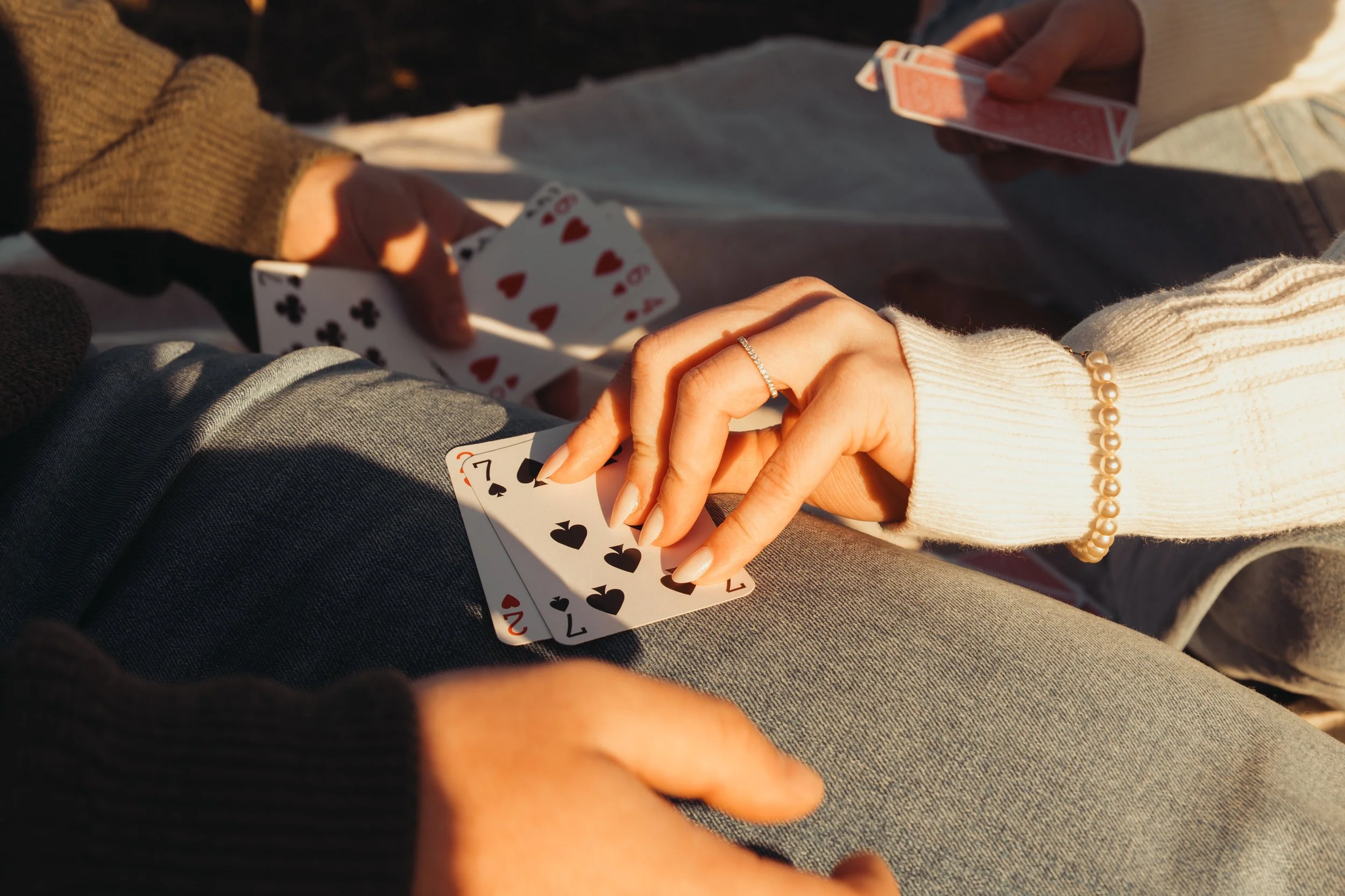 Deux personnes jouent aux cartes, une personne avec un bracelet de perles et une autre portant un bijou à la main. Cartes visibles avec des figures et des chiffres.