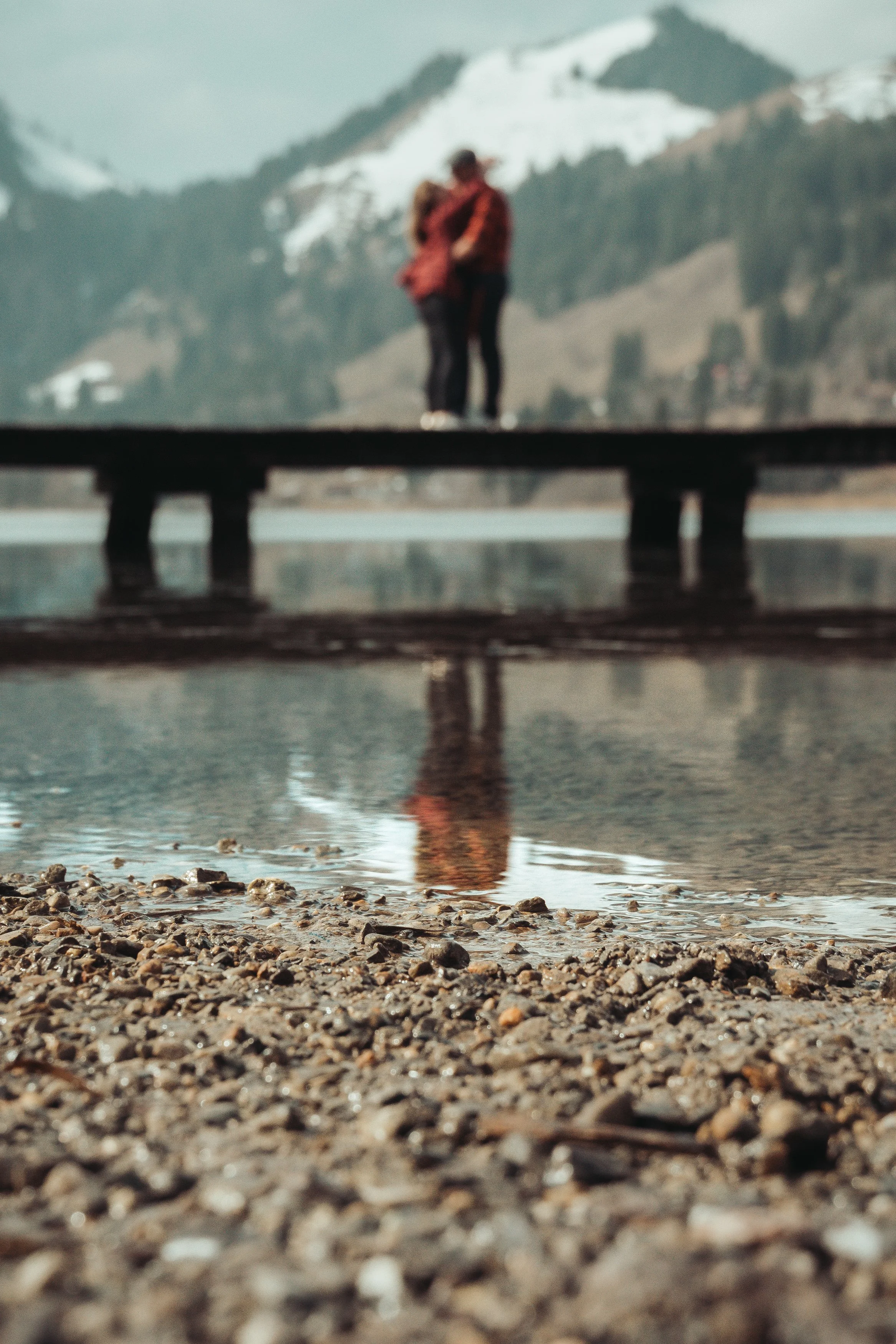 Deux personnes se tiennent sur un pont en bois au-dessus d'un lac, avec des montagnes enneigées en arrière-plan.