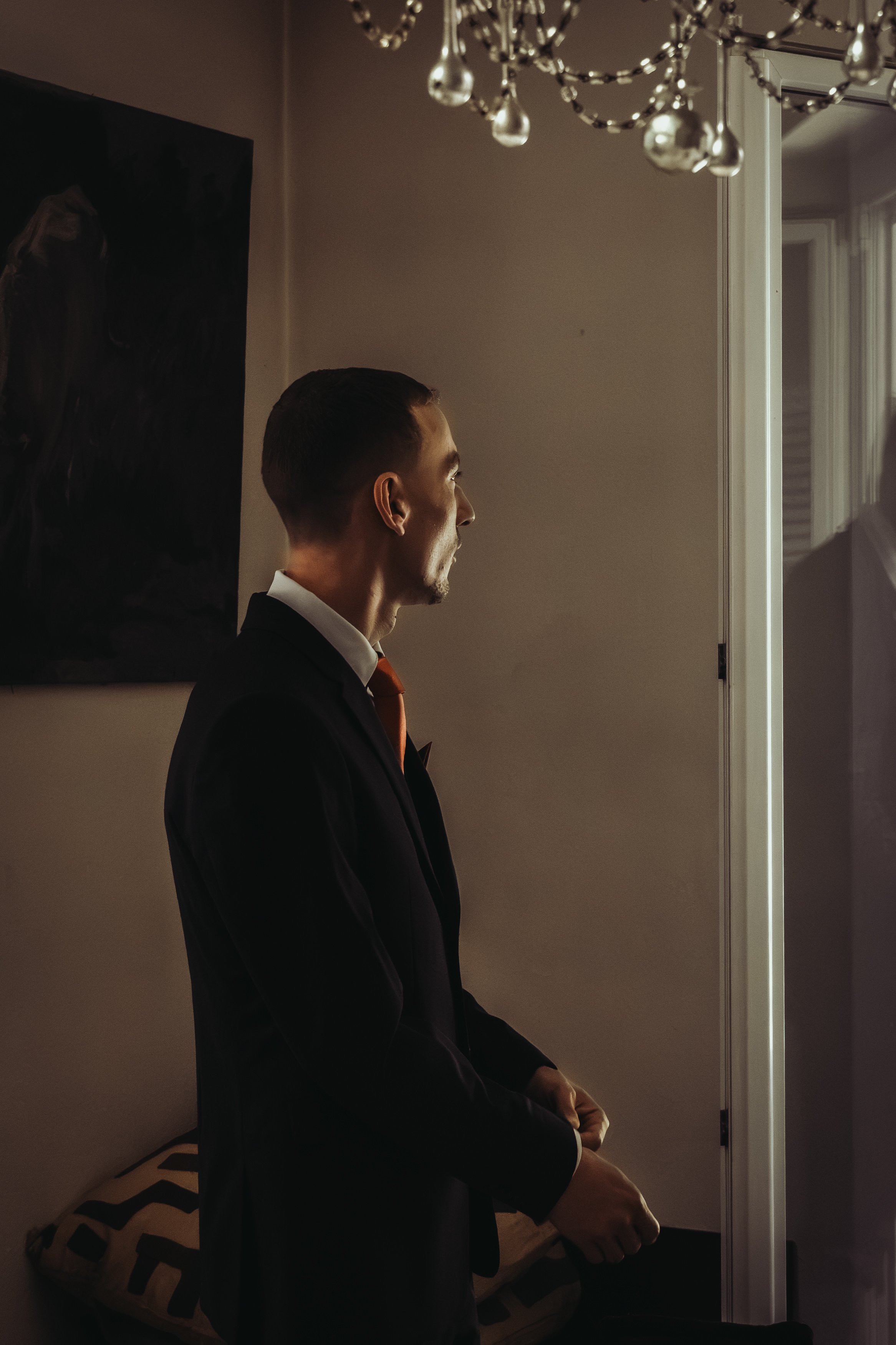 Un homme en costume noir, vu de profil, assis dans une pièce peu éclairée, près d’un miroir et d’un lustre.