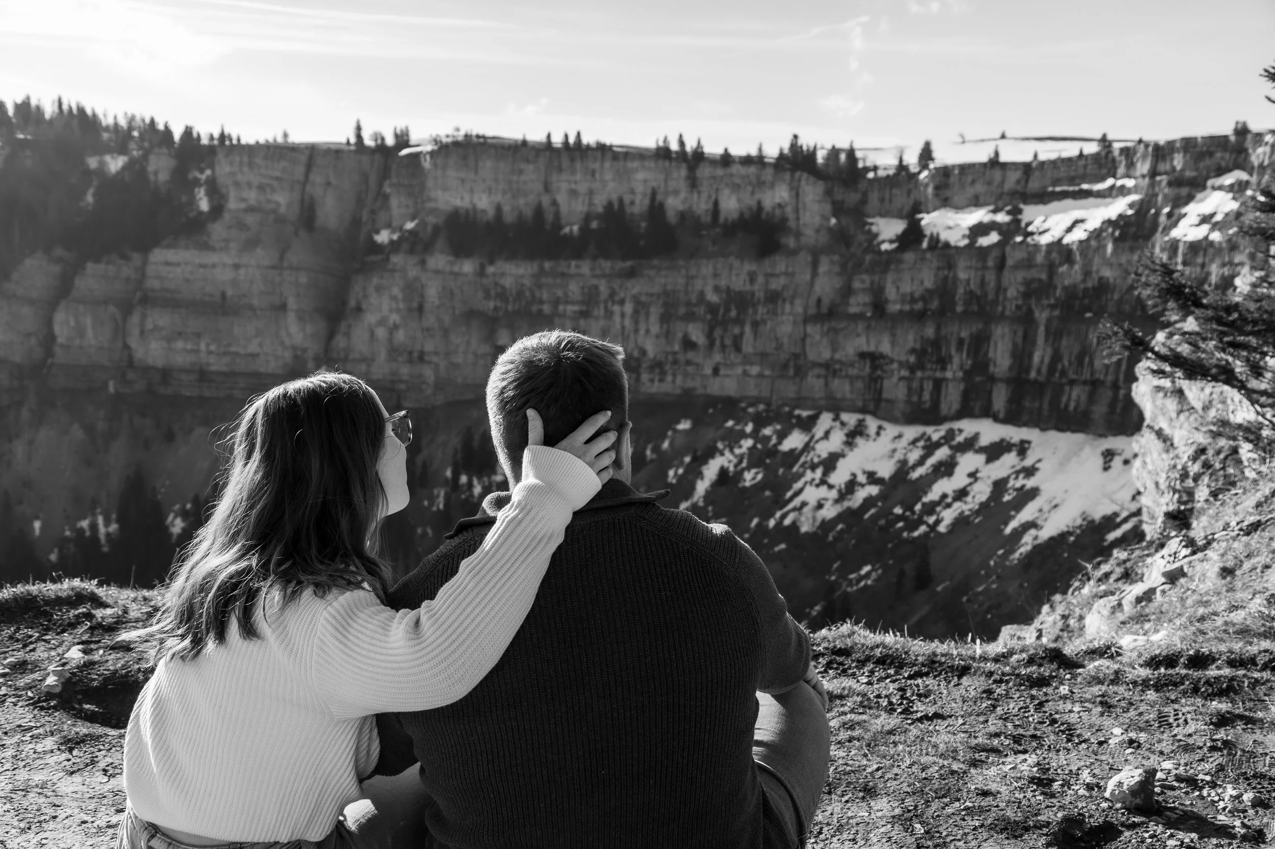 Deux personnes assises près d'un lac, regardant un canyon avec des falaises rocheuses et des arbres, sous un ciel clair, en noir et blanc.