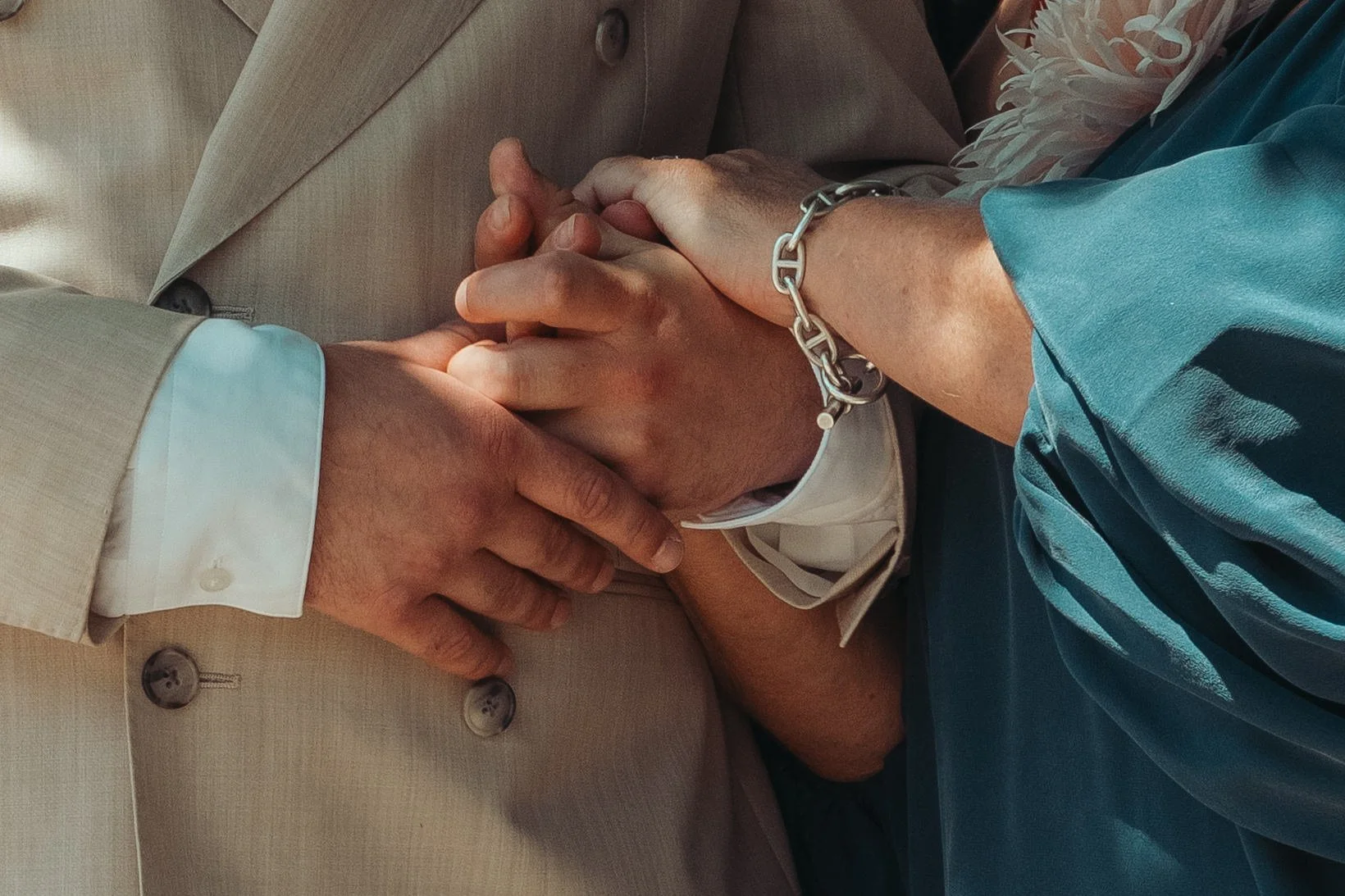 Deux personnes tiennent leurs mains jointes, symbolisant le lien ou l'engagement, l'une portant un costume beige et l'autre une chemise turquoise avec un bracelet en métal.