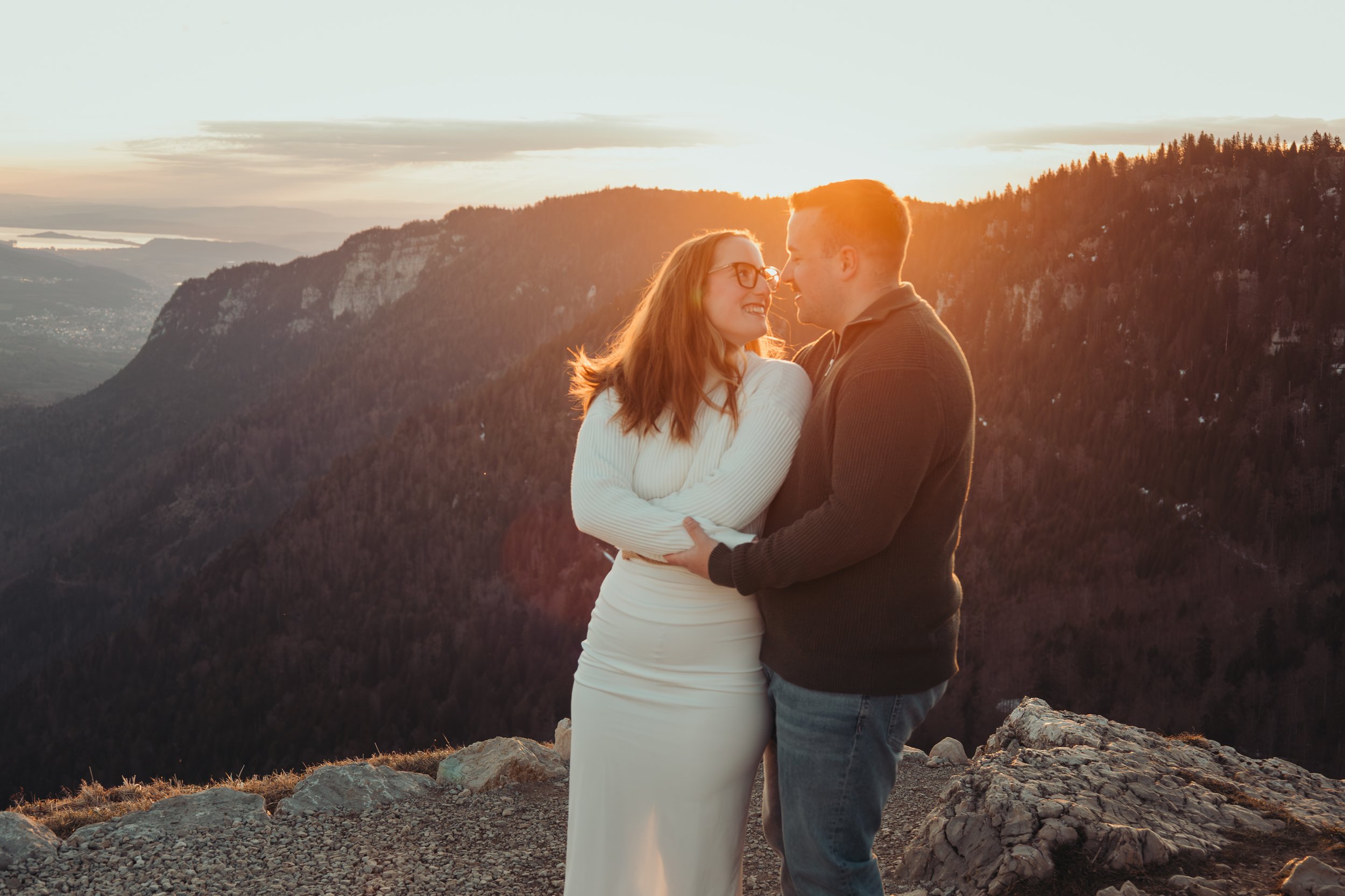 Un couple se tient sur une falaise au coucher du soleil, entouré de montagnes et de paysages naturels.