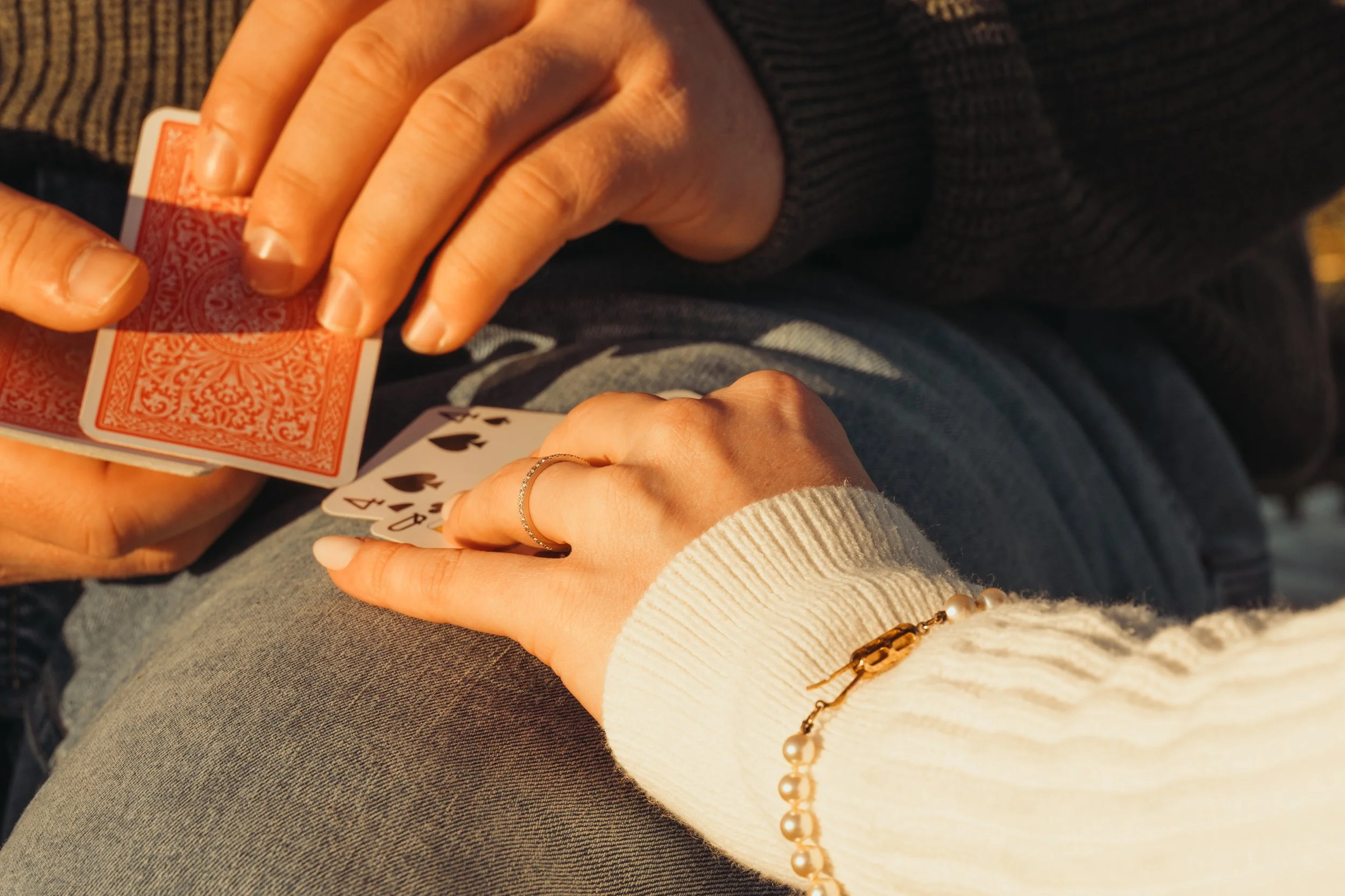 Deux mains tiennent un jeu de cartes, l'une dévoilée et l'autre encore face cachée. La personne porte un bracelet de perles et une bague. La scène semble se dérouler sur les genoux.