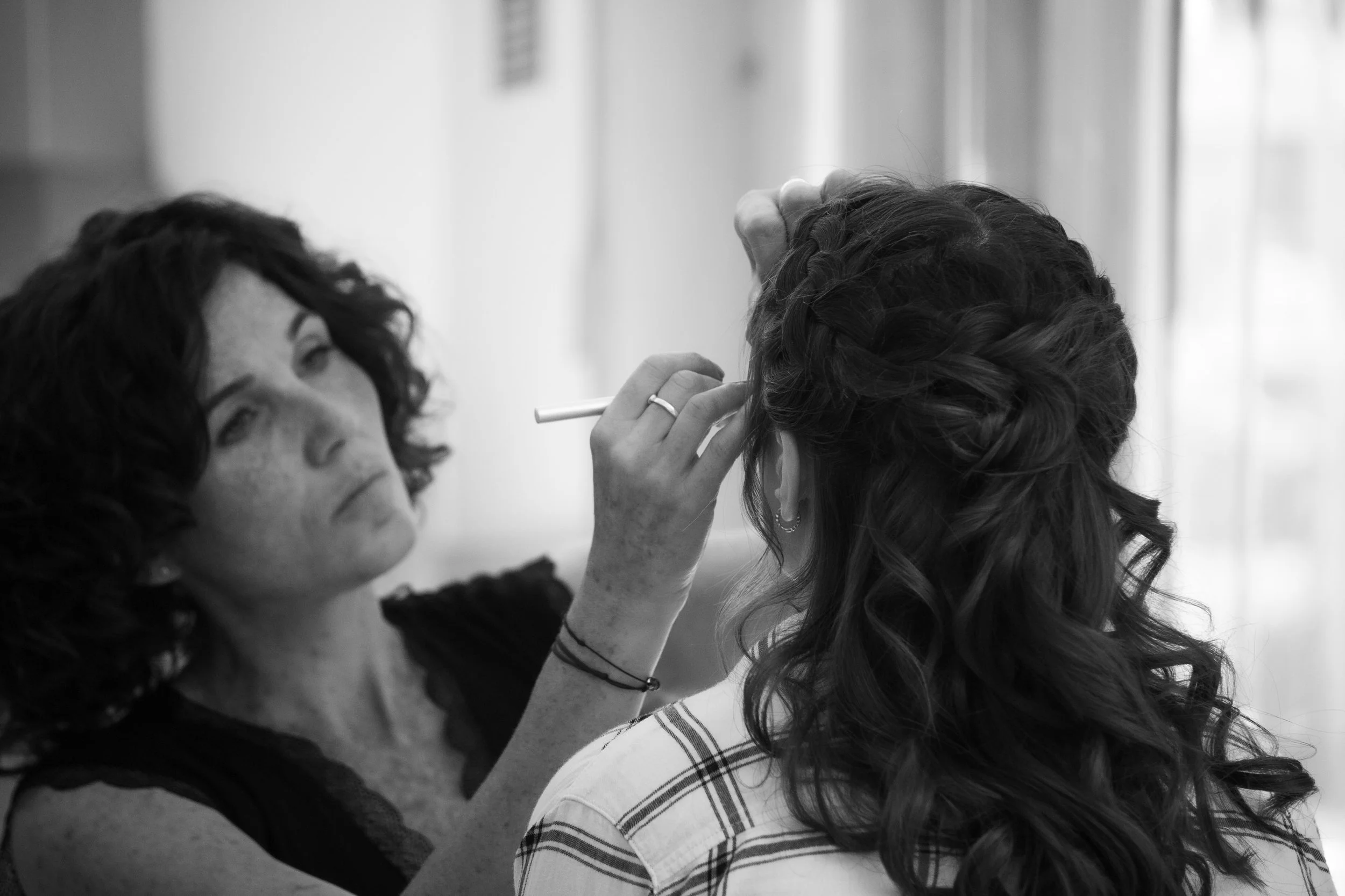 Une femme avec des cheveux bouclés, coiffée en tresse, reçoit un soin de maquillage ou un tatouage temporaire par une autre femme dans un environnement intérieur.