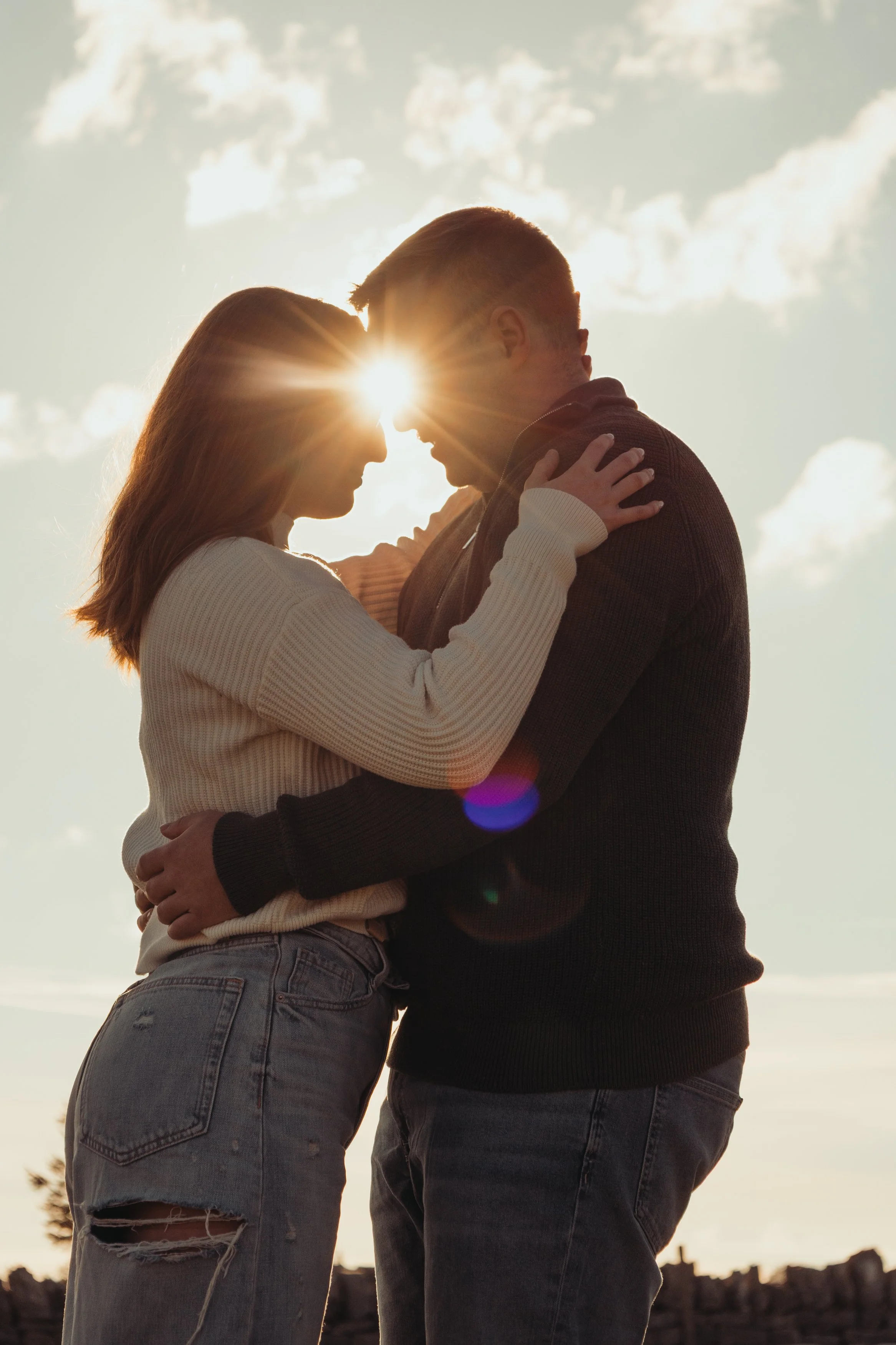 Un couple en chevauchement, regardant chacun l'autre lors du coucher de soleil, avec le soleil derrière leur tête, un ciel partiellement nuageux.